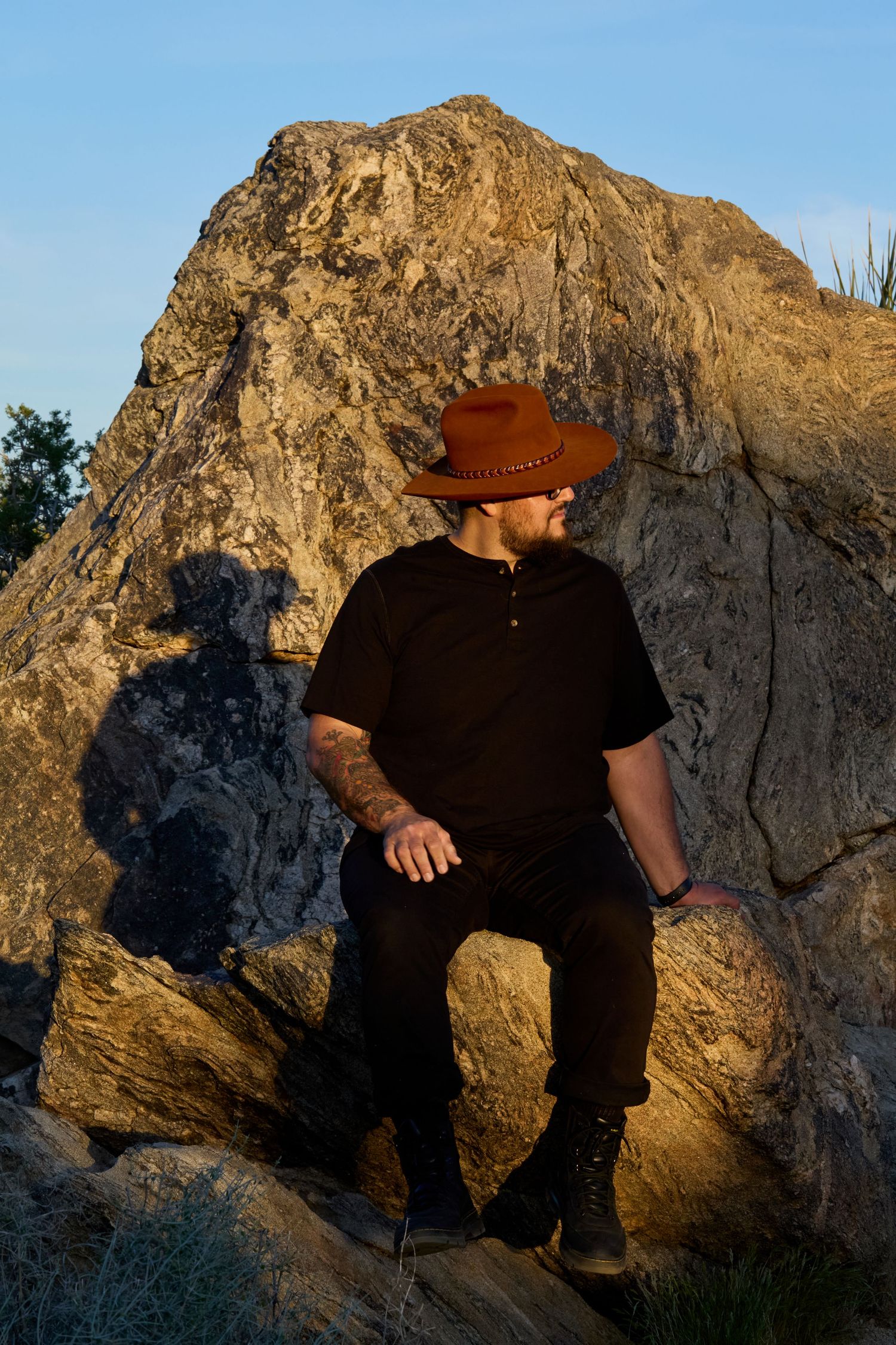 Man in brown cowboy hat and black outfit sitting on rocks with large boulder and blue sky background.