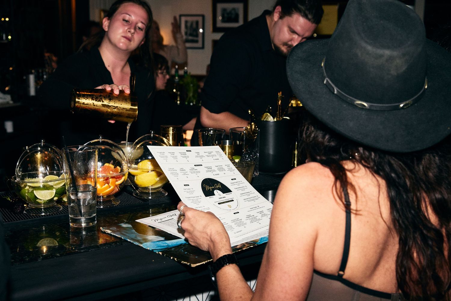 People looking at menus and drinks at a dimly lit bar with cocktail glasses visible.