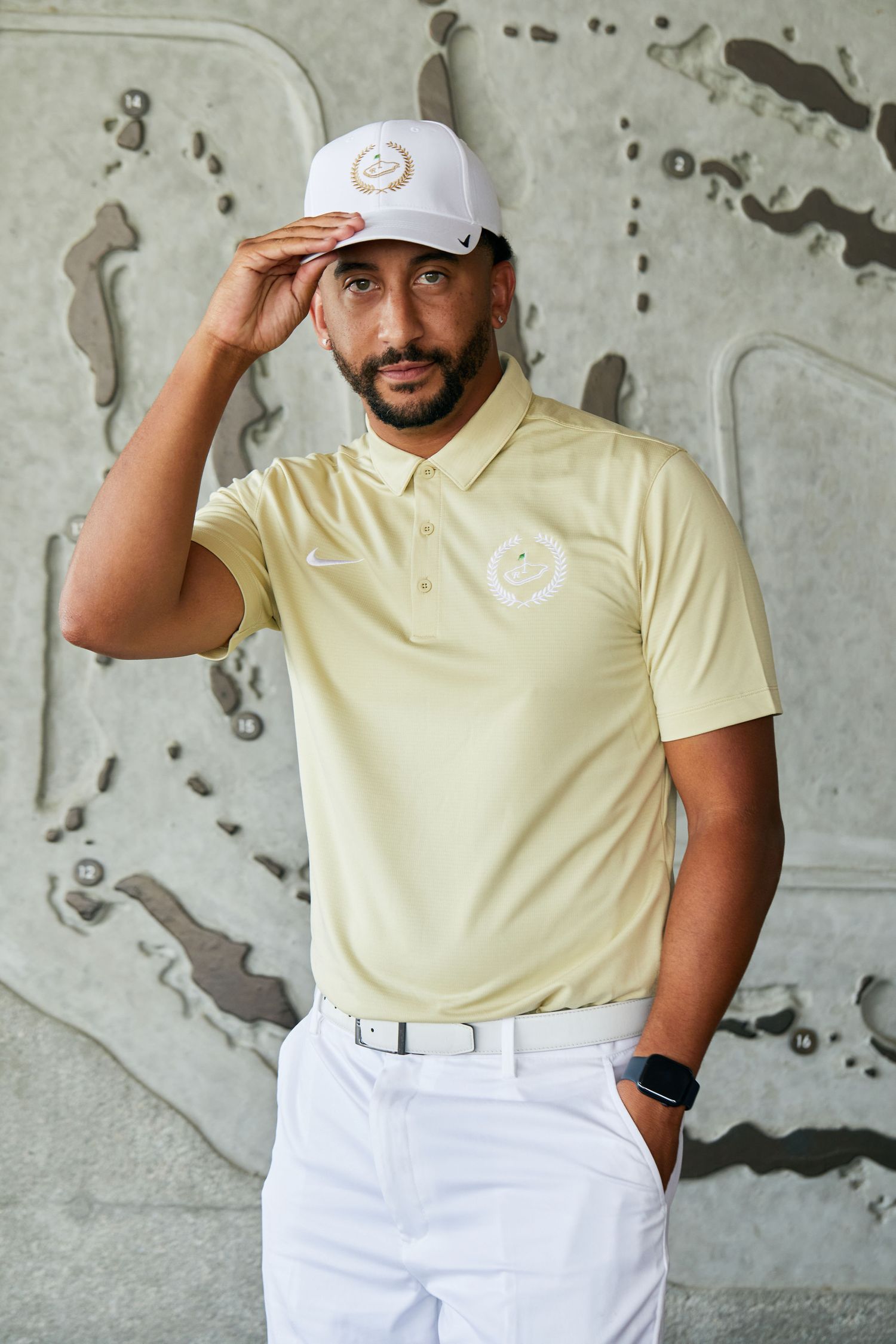Golf attire featuring a pale yellow polo shirt, white cap, and white pants against a patterned wall.