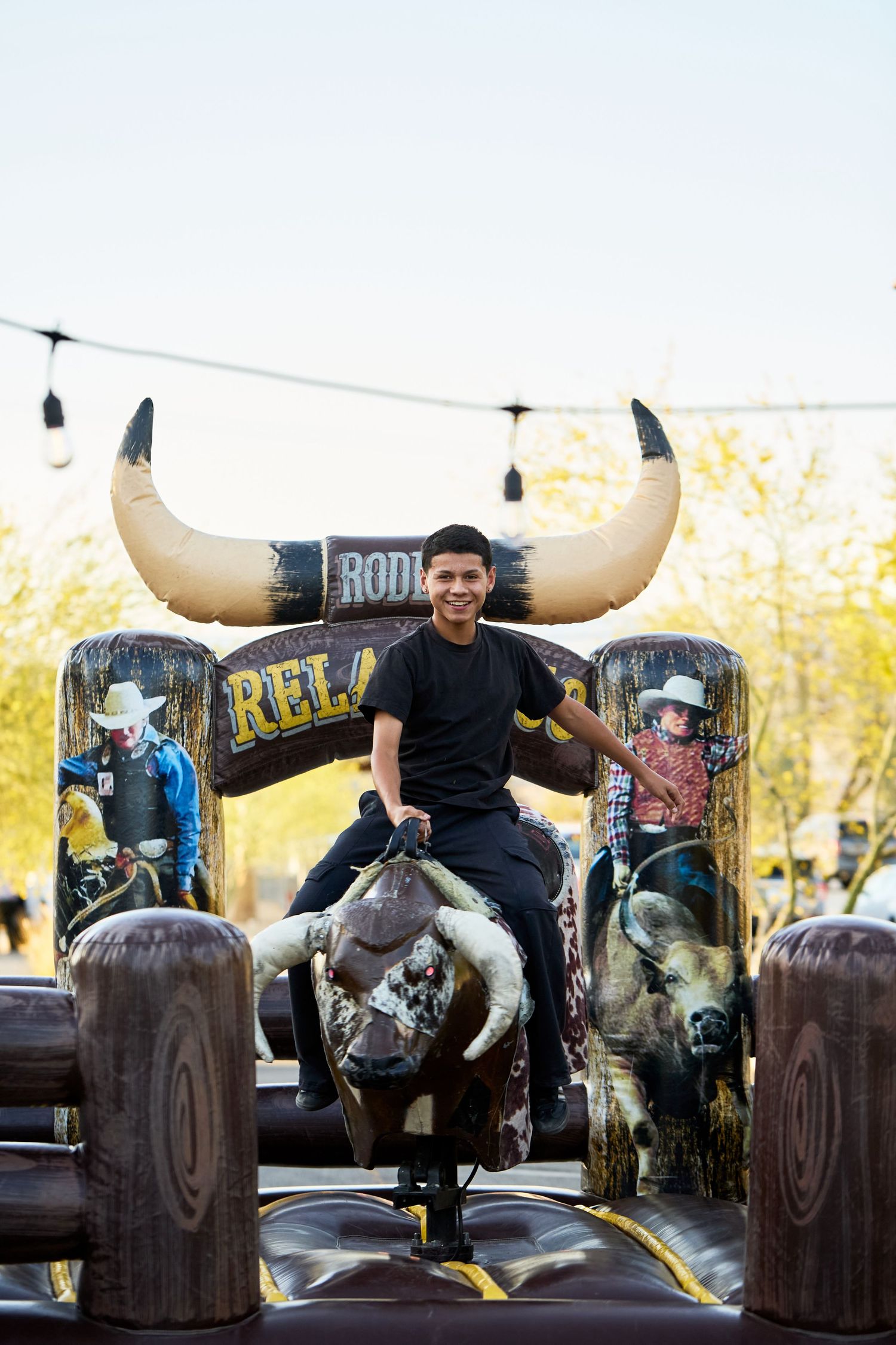 Young man smiling while riding a mechanical bull at an outdoor rodeo-themed event with large inflatable horns.