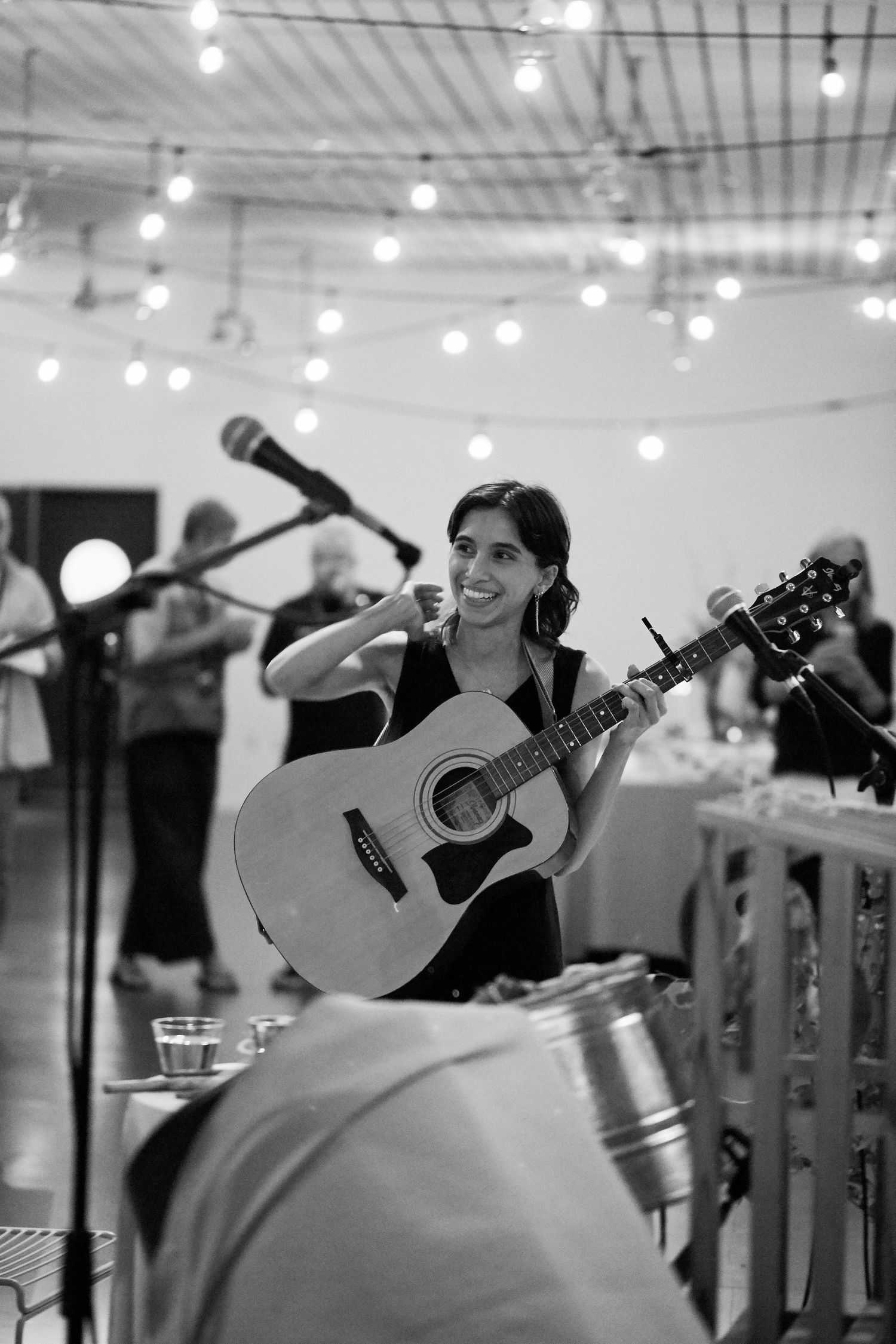 A musician performs with an acoustic guitar under string lights at an indoor venue in black and white.