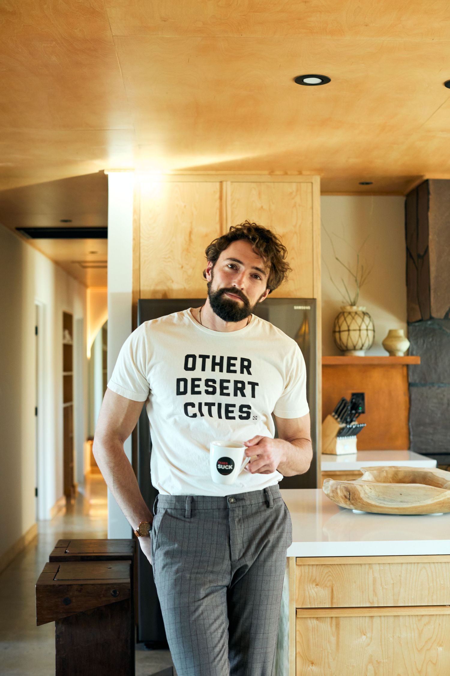 Person in grey pants and white t-shirt with 'DESERT CITIES' text stands in modern minimalist interior space.