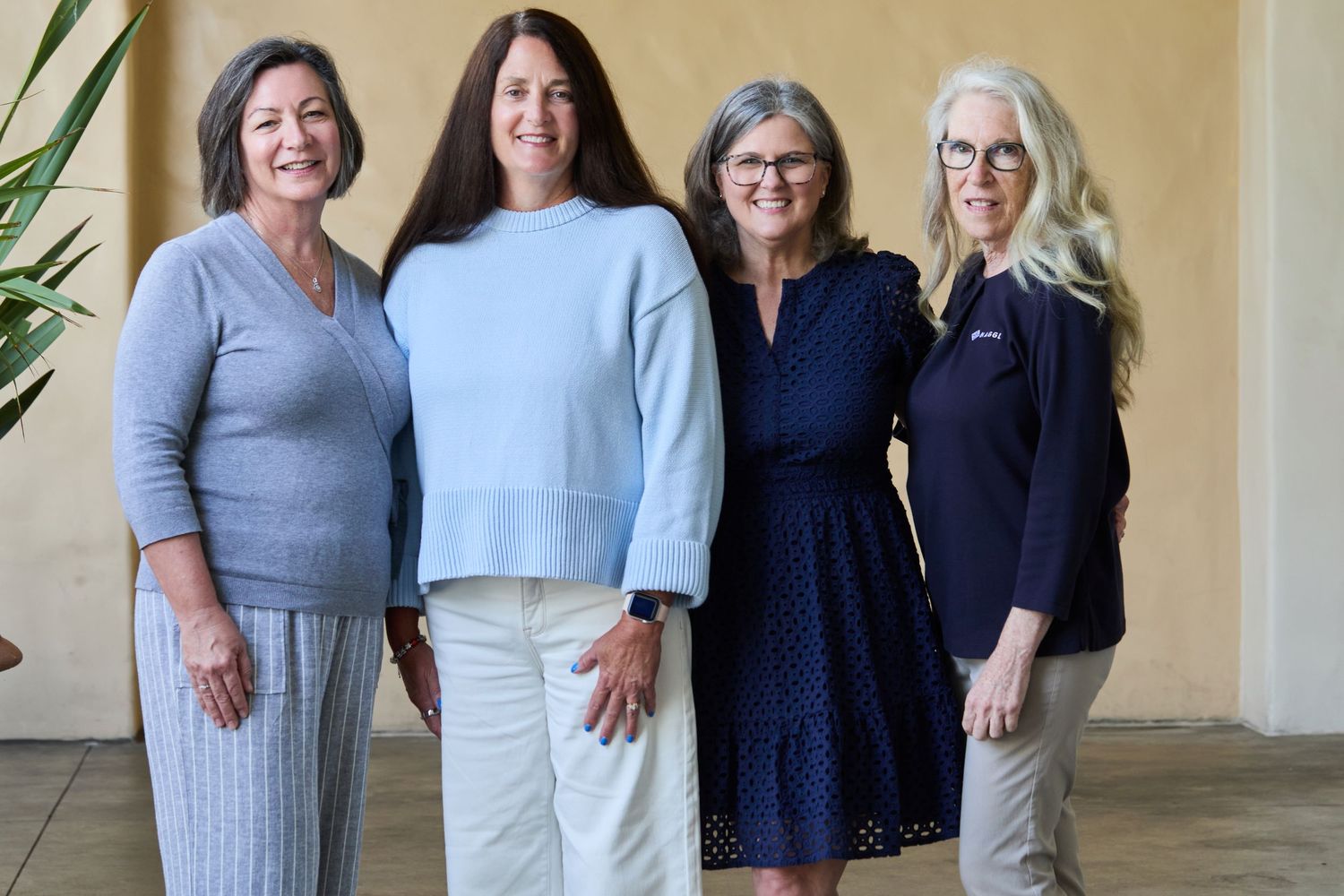 Four women stand together wearing casual business attire in shades of blue, gray and white against a beige wall.