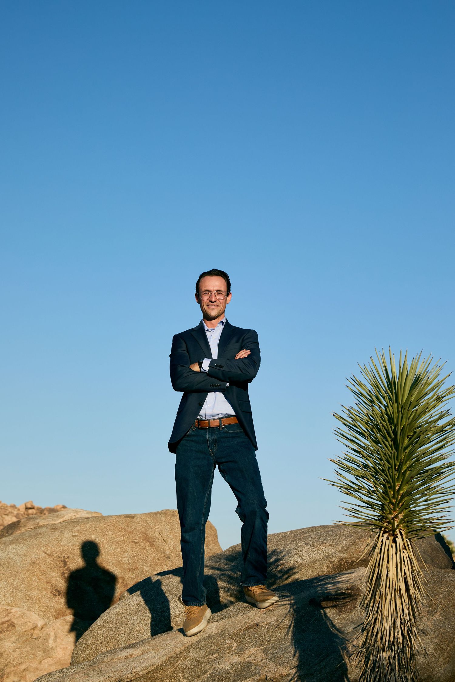 A figure in a dark suit stands confidently against a desert landscape with yucca plants and clear blue sky.