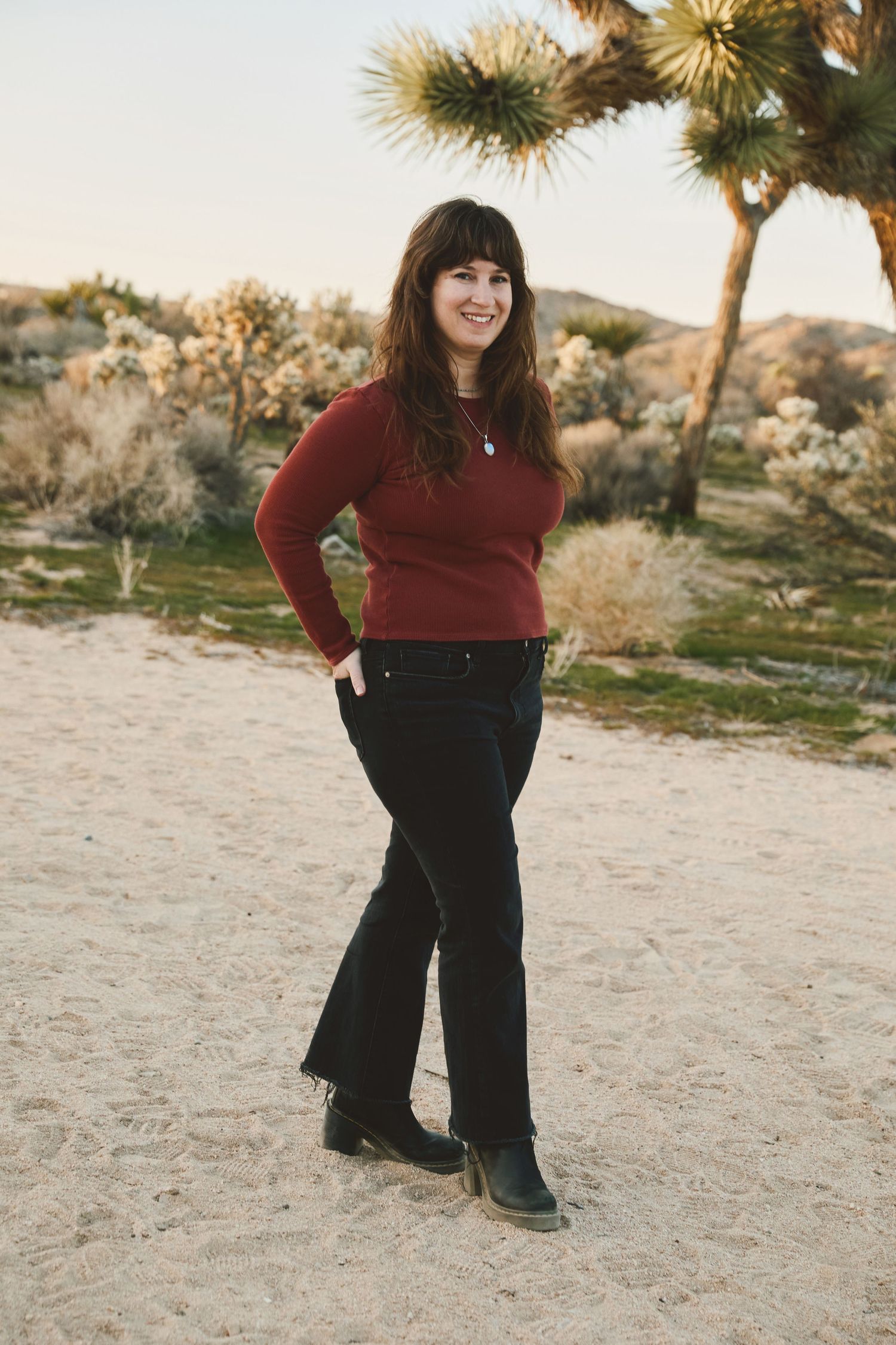 A person in a burgundy sweater and black pants poses outdoors in a desert landscape with Joshua trees at sunset.