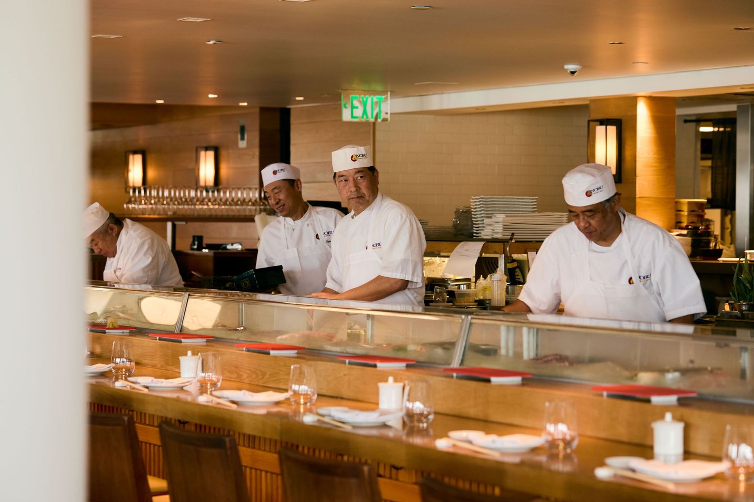 Chefs in white uniforms working behind an elegant sushi bar counter with warm lighting and place settings.