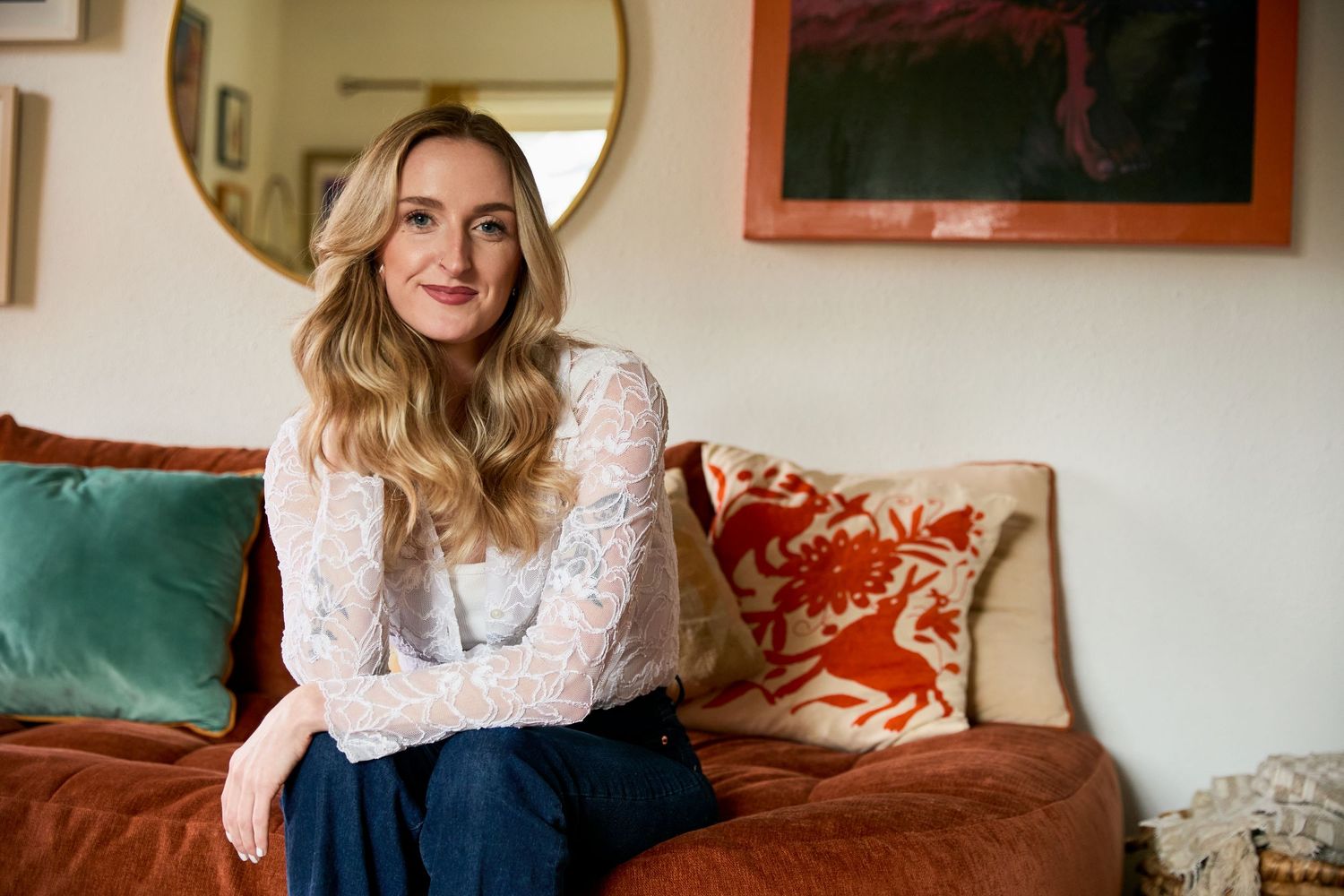 Woman with blonde wavy hair in white lace top sits on rust velvet sofa with colorful pillows in stylish home.