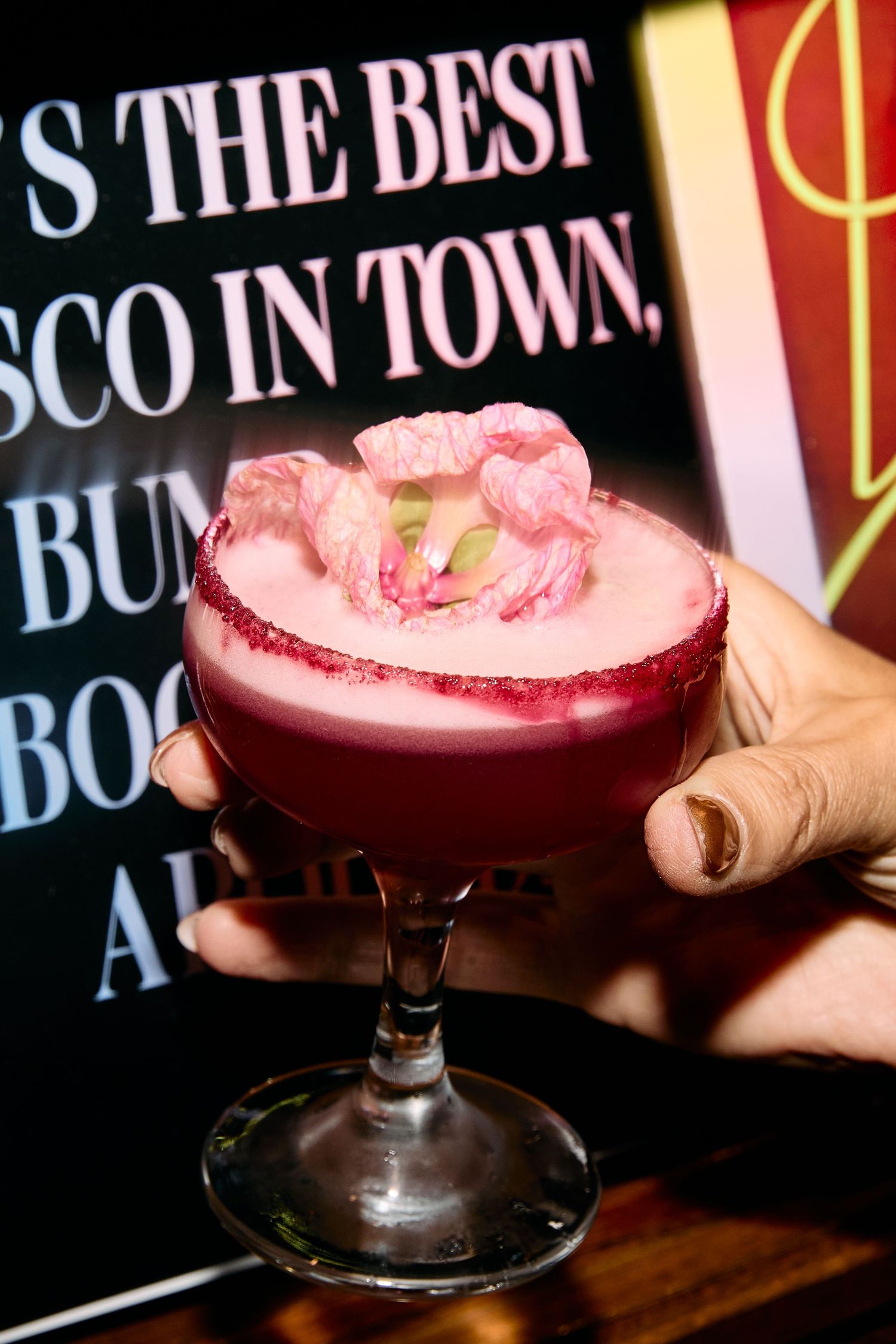 A pink craft cocktail with a sugar rim and rose garnish is held up against a neon-lit bar sign.