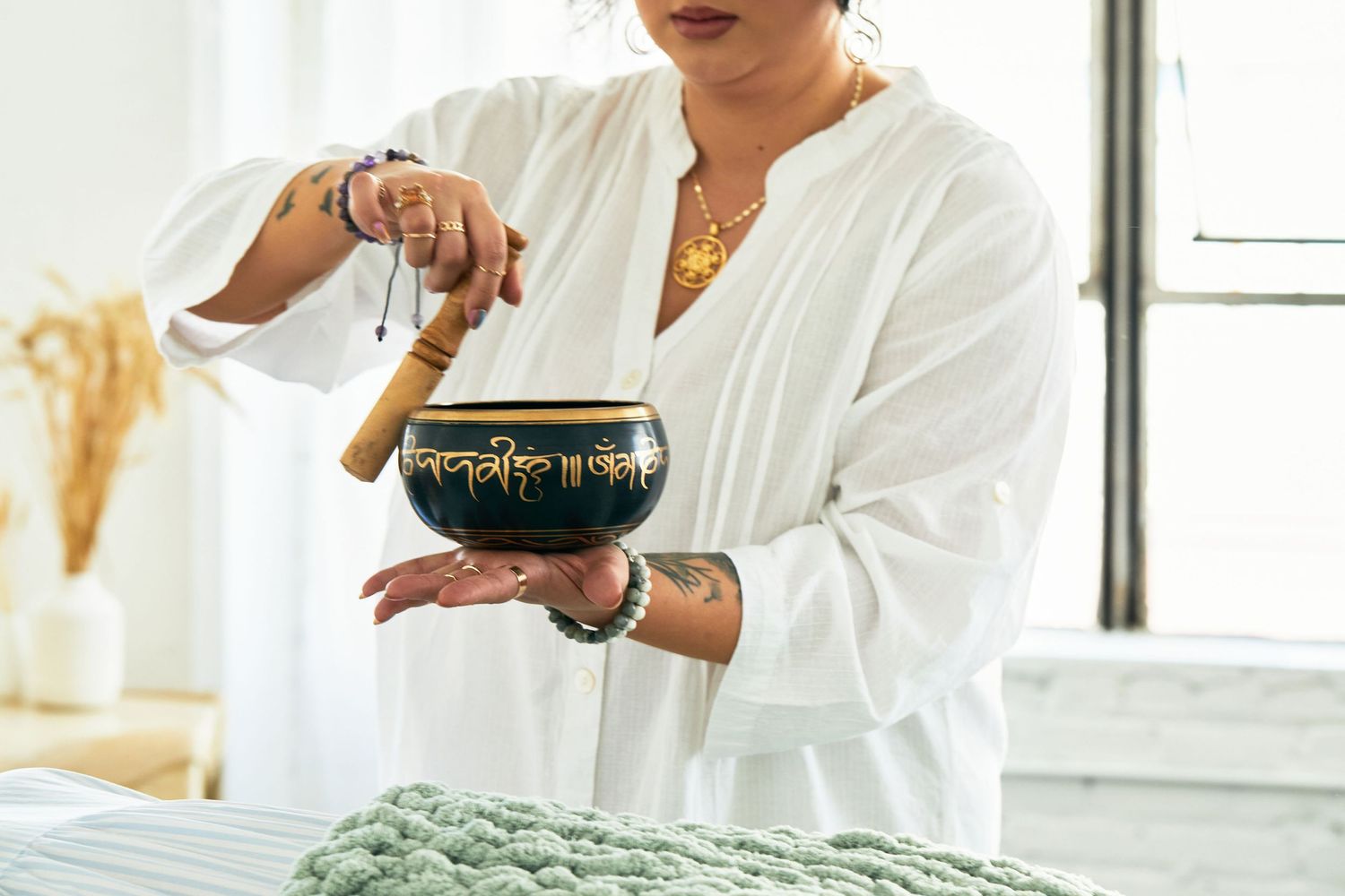 Woman in white robe holding a Tibetan singing bowl with a wooden mallet during a sound healing session.