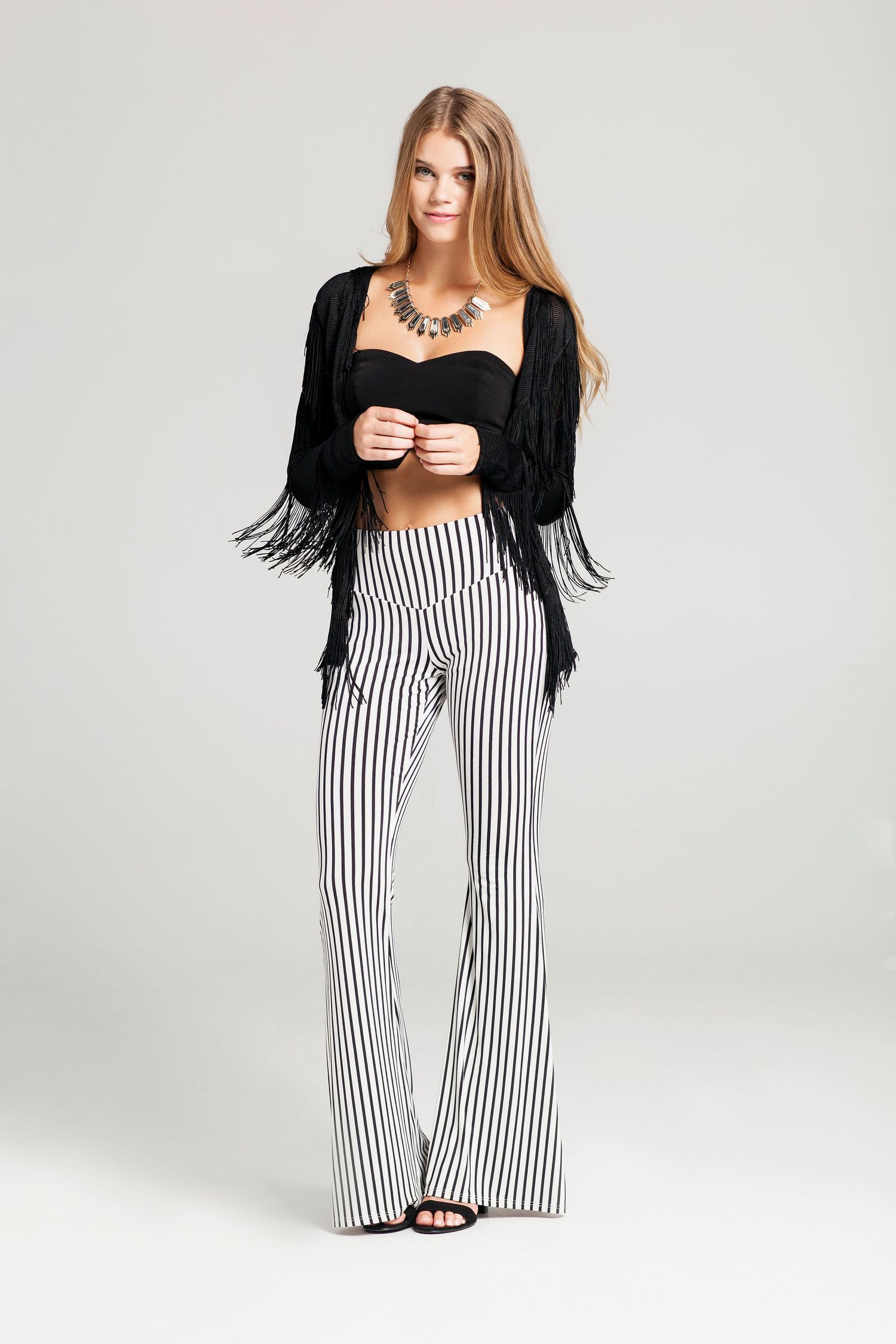 Fashion model wearing black fringe cardigan with striped wide-leg pants and crop top against grey background.