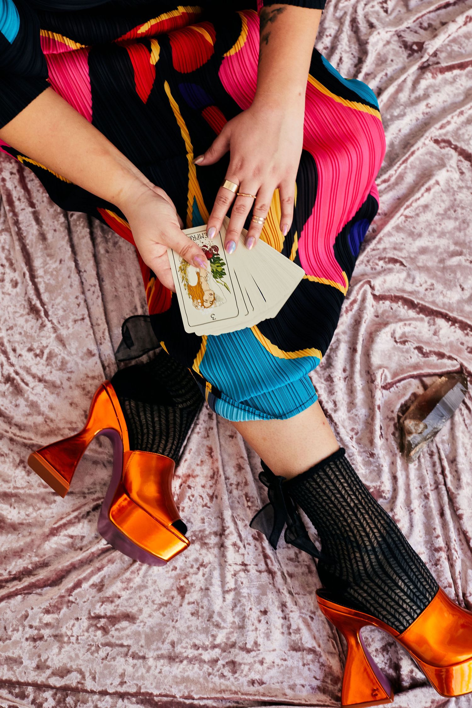 Person sitting on velvet fabric holding tarot cards, wearing colorful dress, black fishnet socks, and orange metallic platform heels.