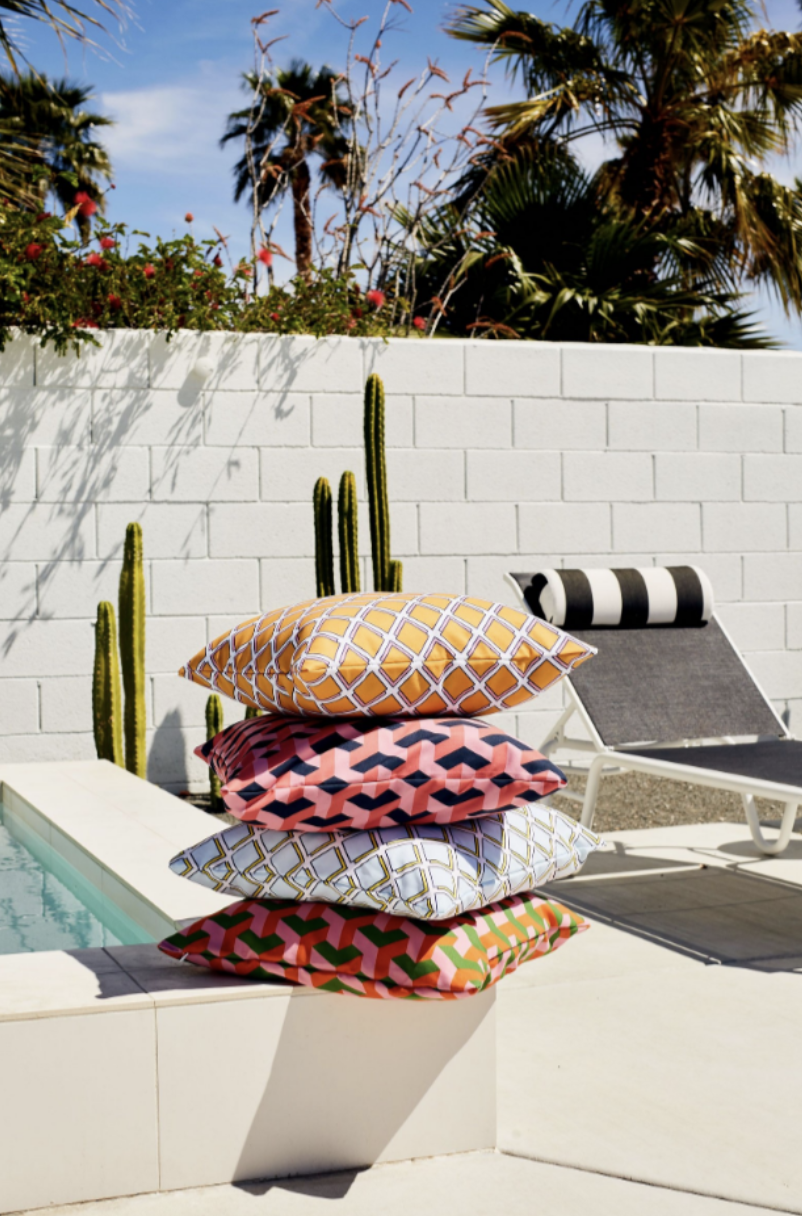 Stacked colorful geometric pattern pillows by pool with palm trees and white wall.