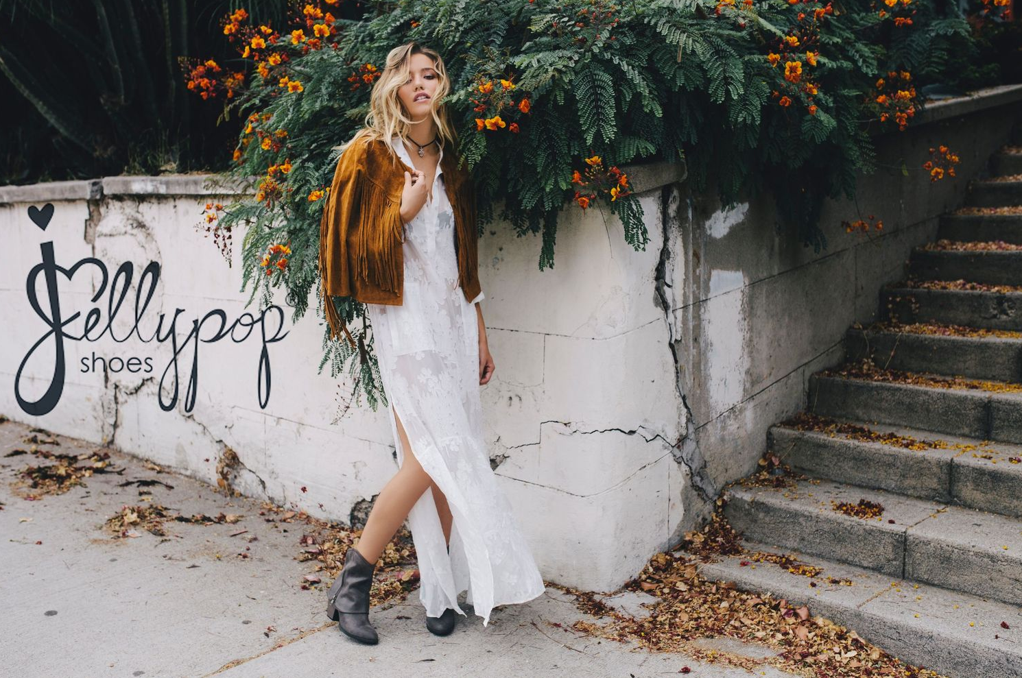 Stylish autumn outfit with brown jacket and white dress photographed against textured wall with plants.