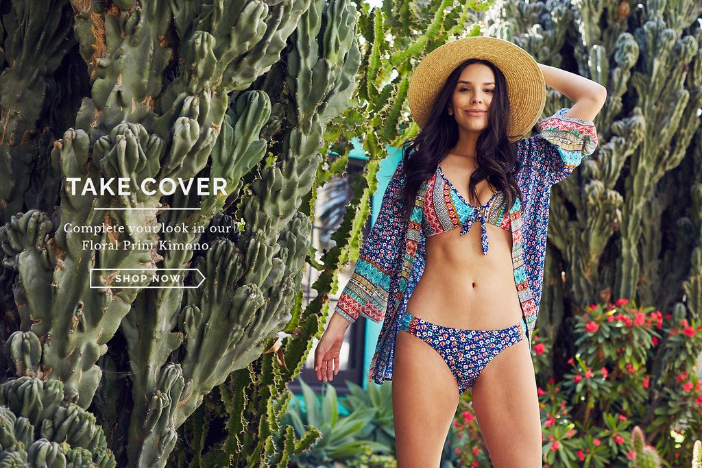 Model in blue patterned swimwear and wide-brimmed hat poses in tropical garden setting.