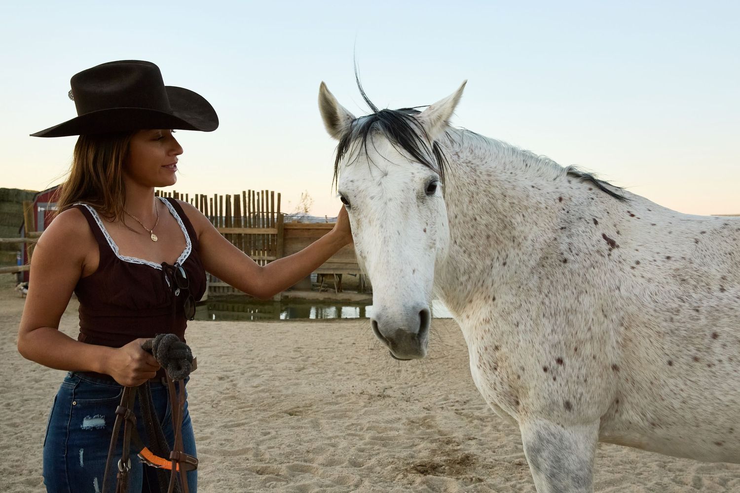 A cowgirl in a black hat gently pets a white speckled horse on a sandy ranch at dusk.