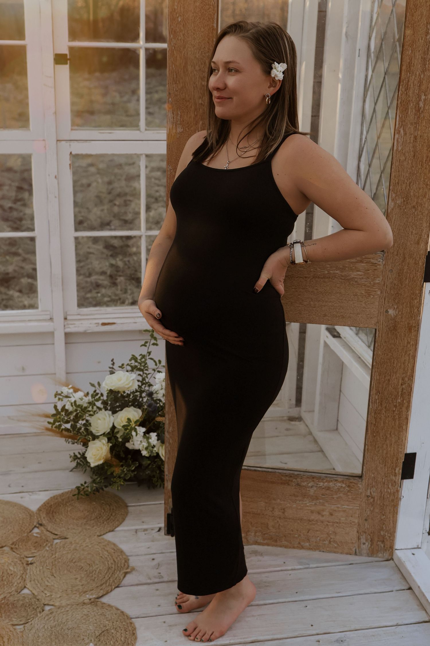 Woman posing in black dress against rustic white walls and windows.