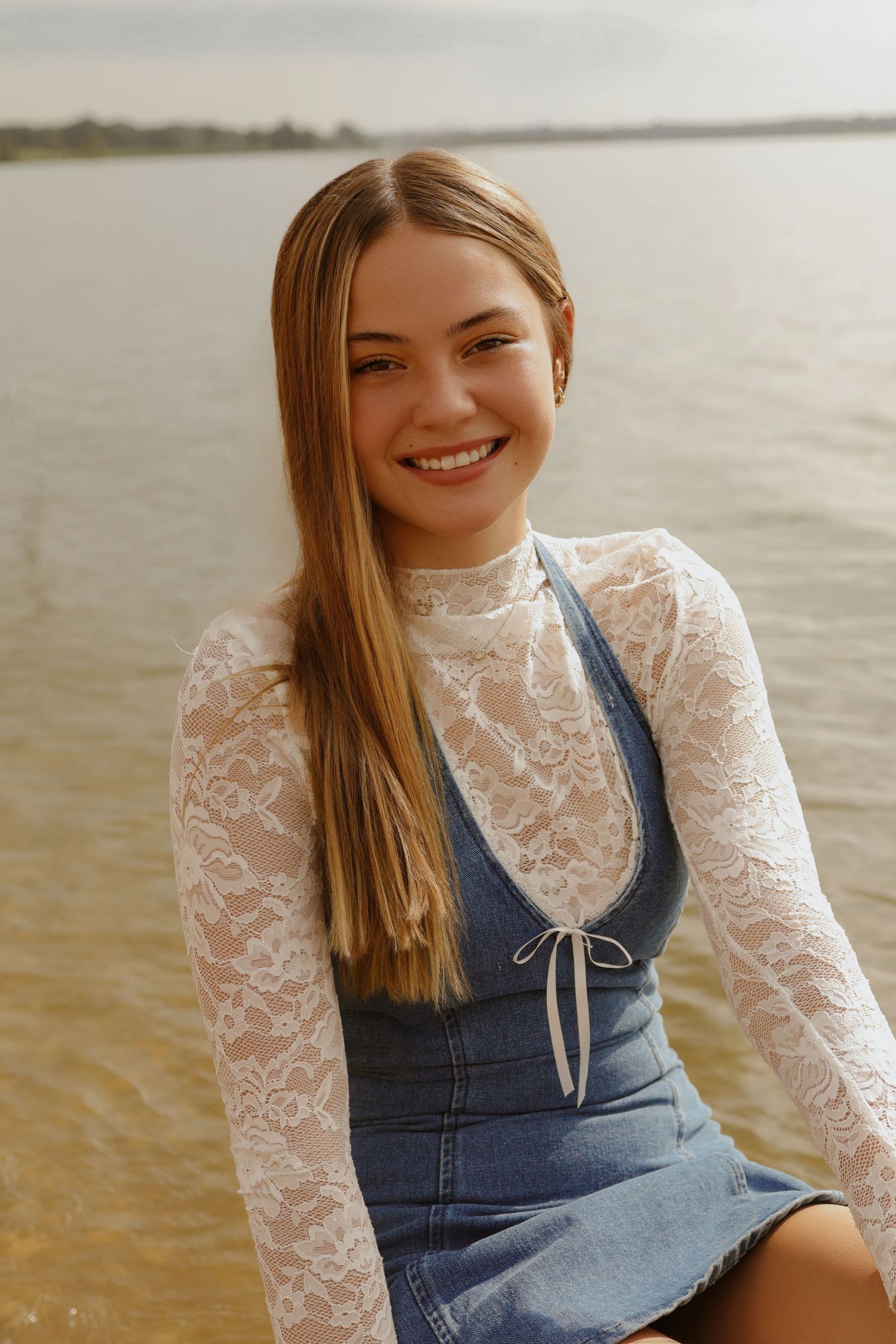Person wearing a white lace top and denim overalls poses by the water at sunset.