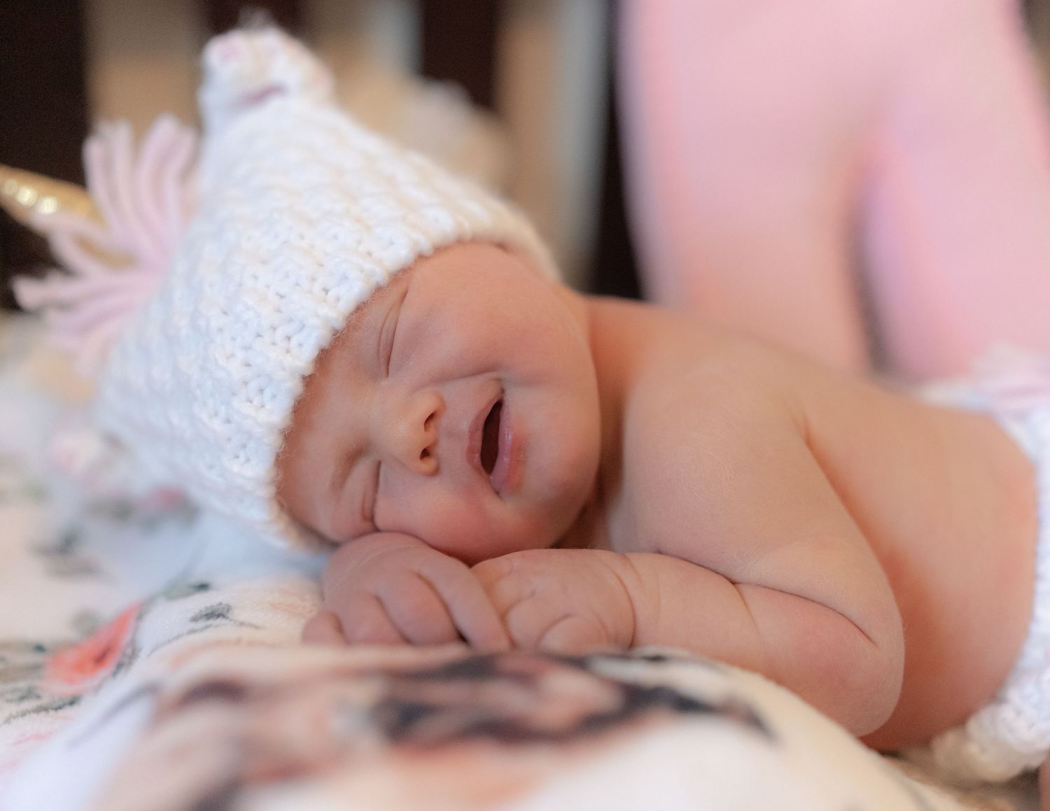 Newborn baby sleeping peacefully wearing a white knit hat, cradled in a parent's arms on a soft floral blanket.