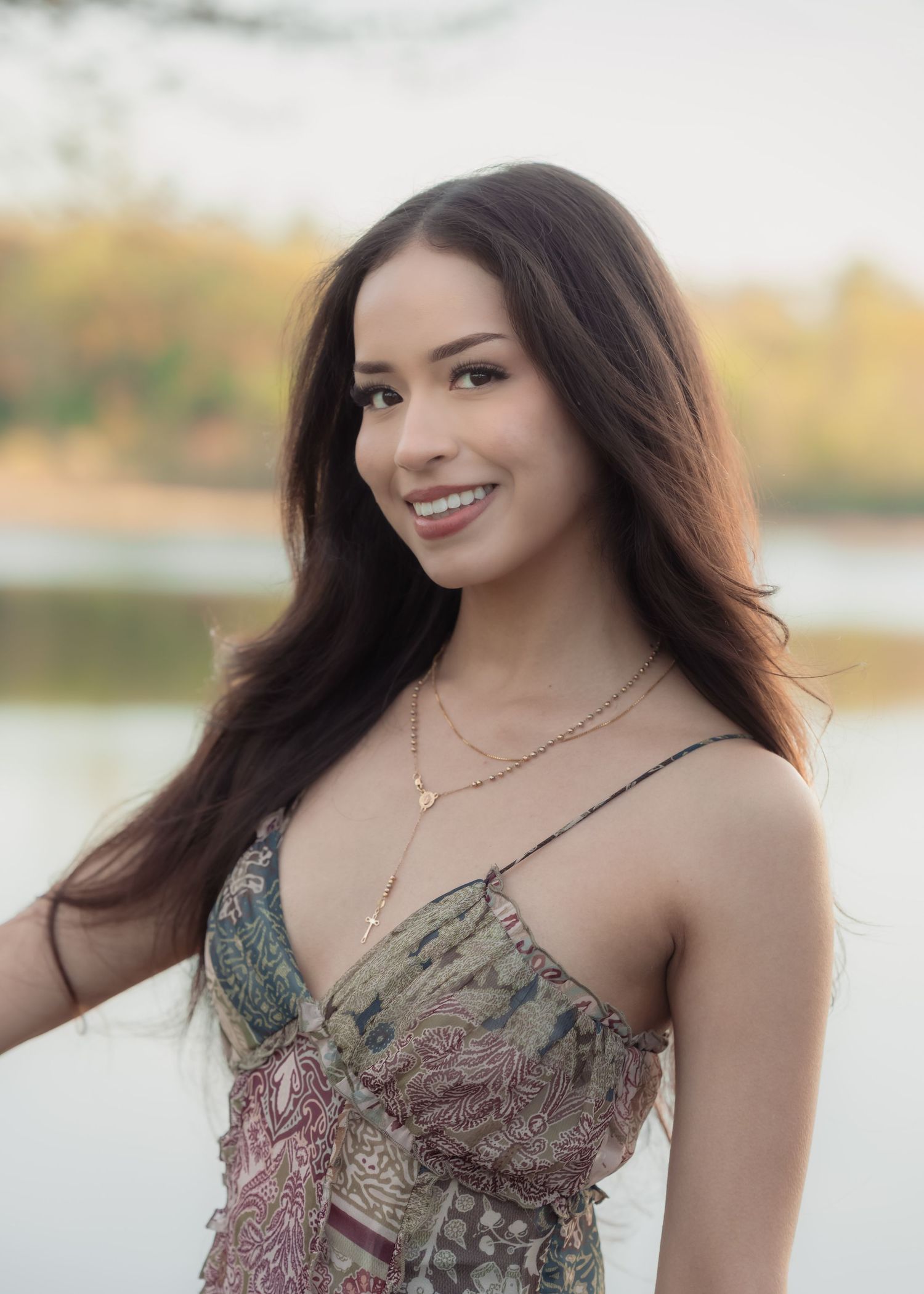 Smiling young woman with long dark hair wearing a floral dress and gold necklaces outdoors near a lake.