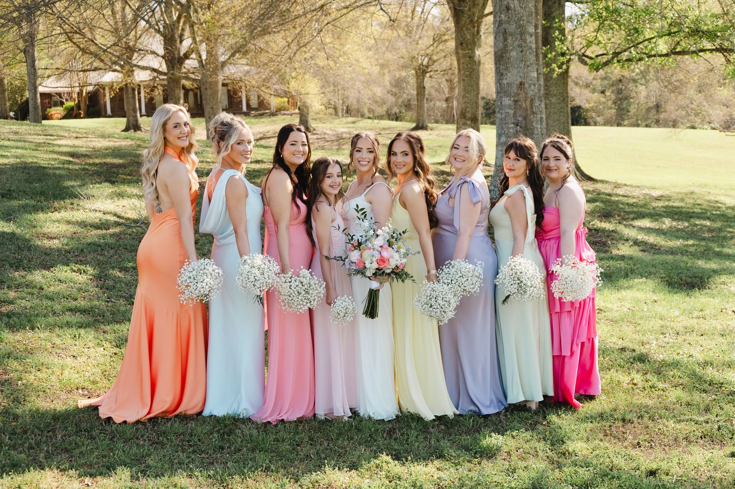 Bridesmaids in rainbow-colored gowns holding white bouquets pose together on green lawn at wedding.