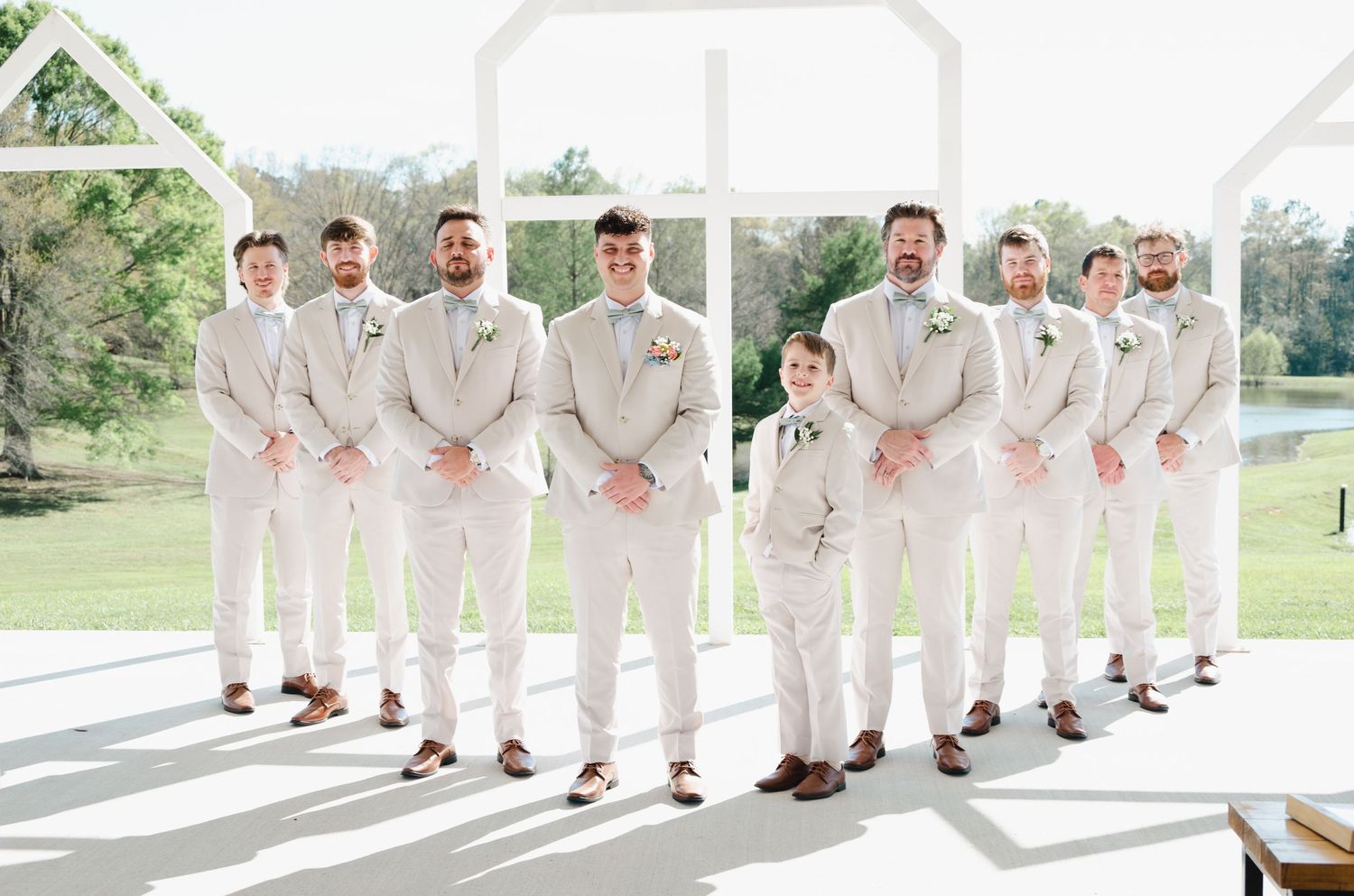 Groom and groomsmen pose in matching tan suits with boutonnieres inside a bright white wedding venue.