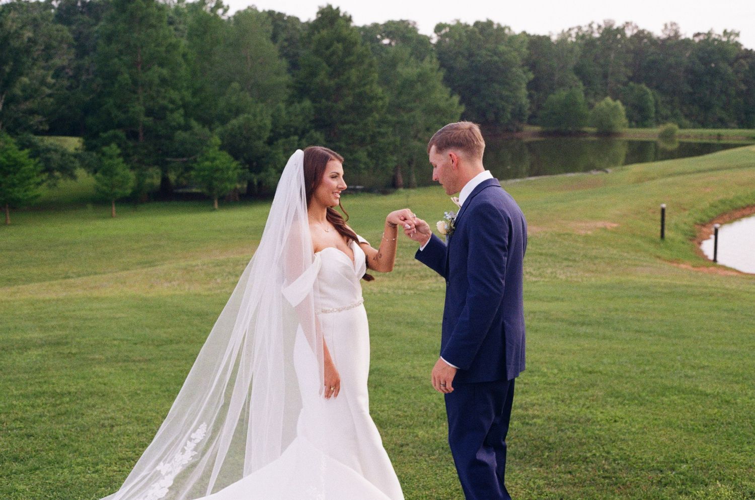 Bride in long veil and groom in navy suit hold hands on a green landscape.