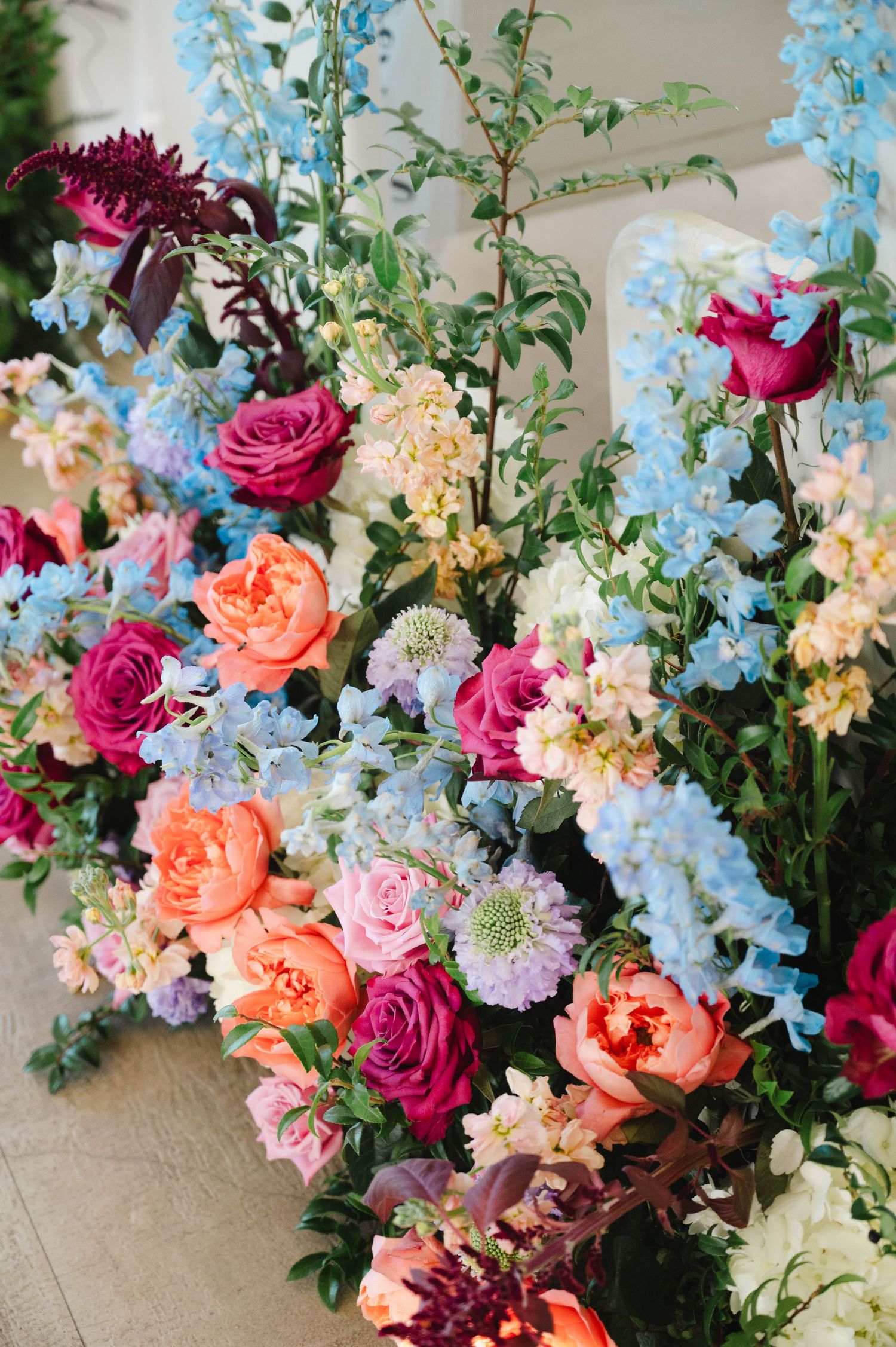 Lush floral arrangement featuring pink roses, orange peonies, blue delphiniums, and burgundy blooms for a wedding ceremony.