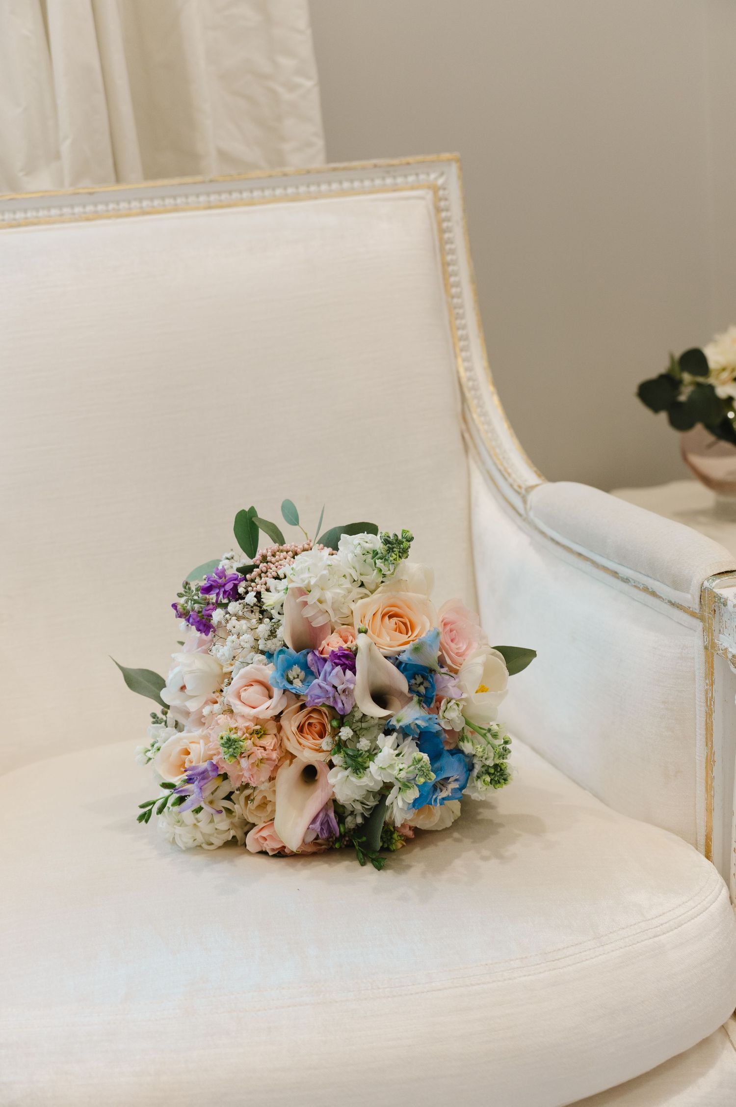 Elegant bridal bouquet with pastel roses, blue flowers, calla lilies, and greenery resting on a white upholstered chair.