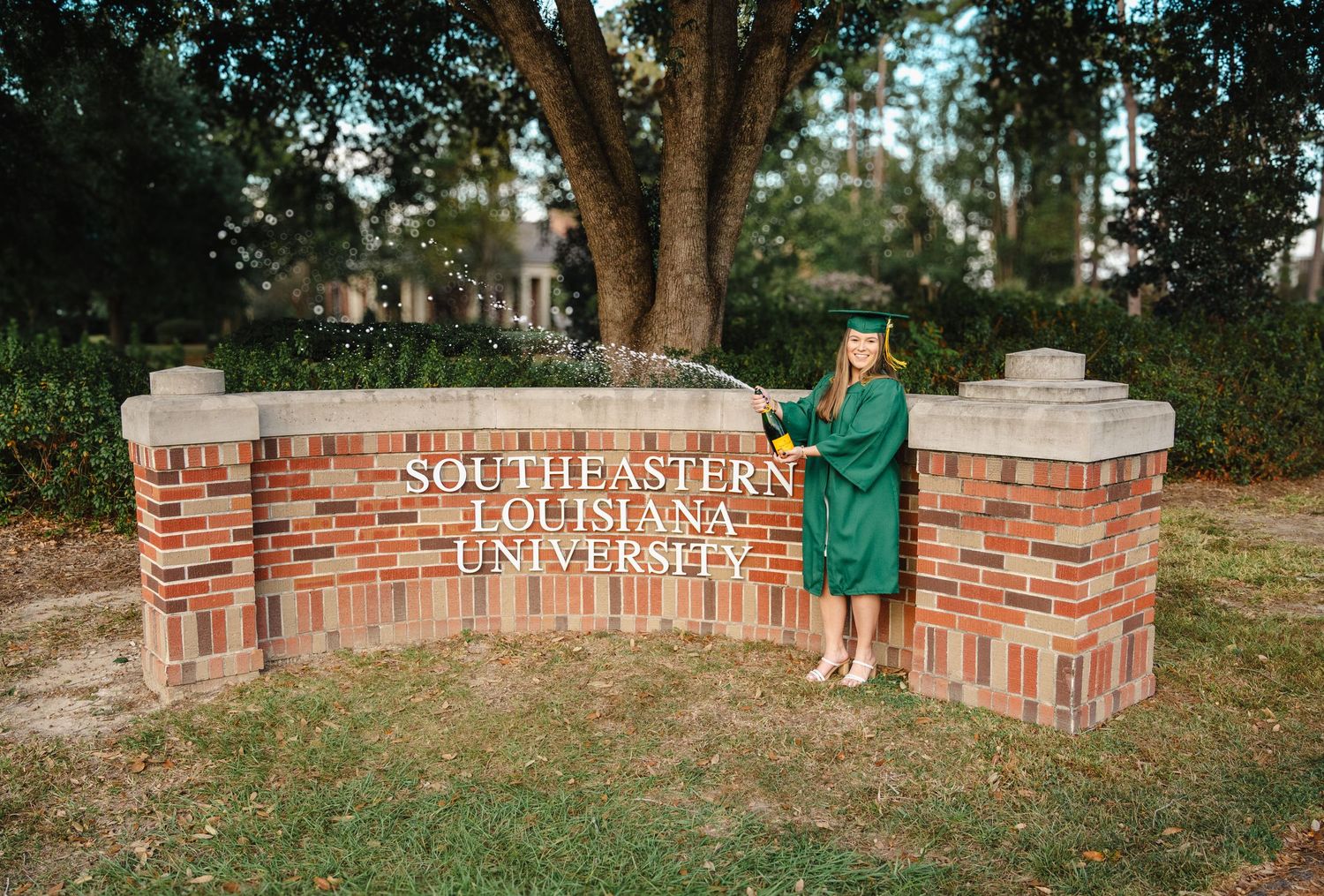 Graduate in green cap and gown poses by Southeastern Louisiana University brick sign outdoors.