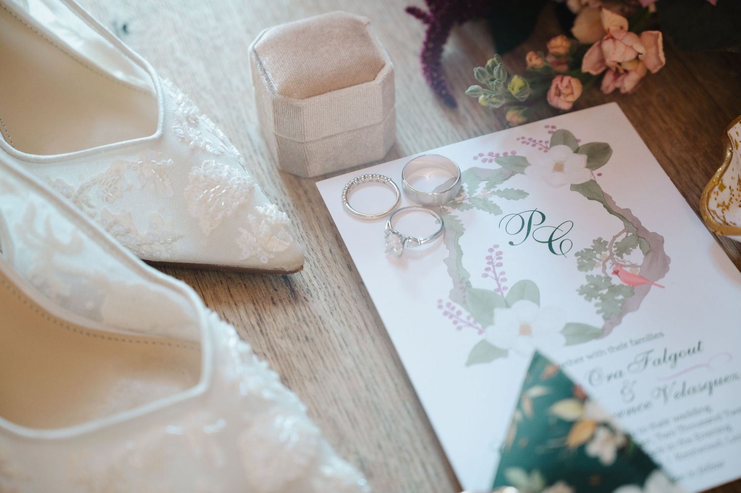 Elegant wedding flat lay featuring lace bridal heels, gold rings, a floral invitation, and a jewelry box on a wooden surface.
