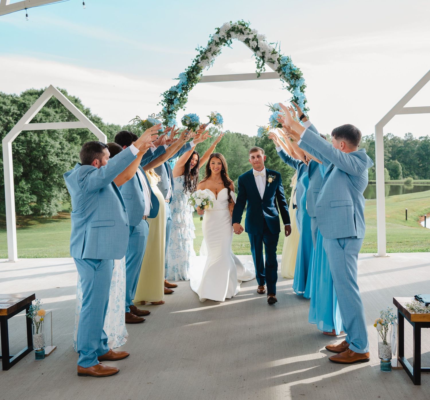 Bride and groom walk through an arch of raised hands at an outdoor wedding ceremony with blue floral decor.