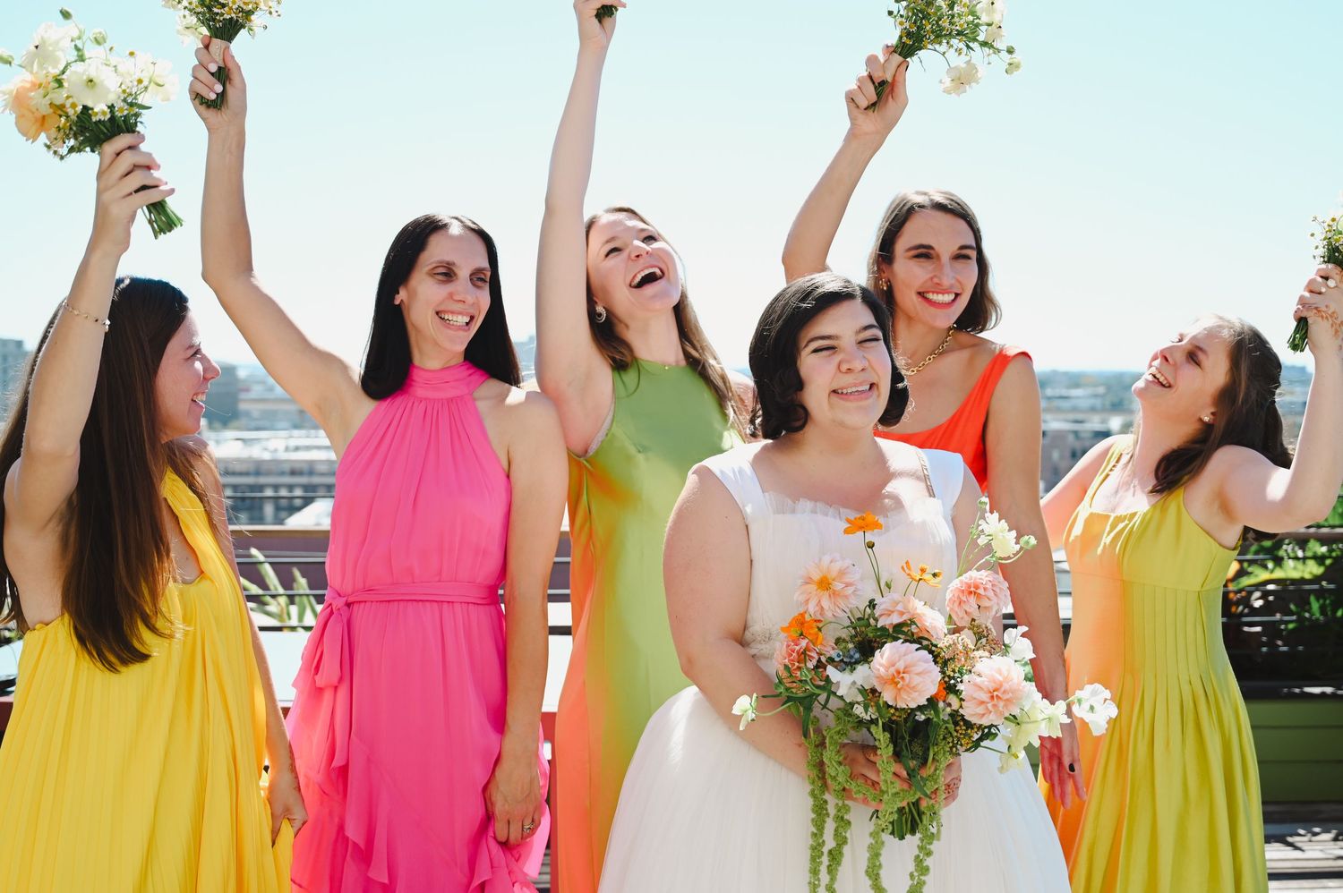 Bride with bridesmaids in colorful yellow, pink, green, and orange dresses celebrating outdoors with bouquets raised.