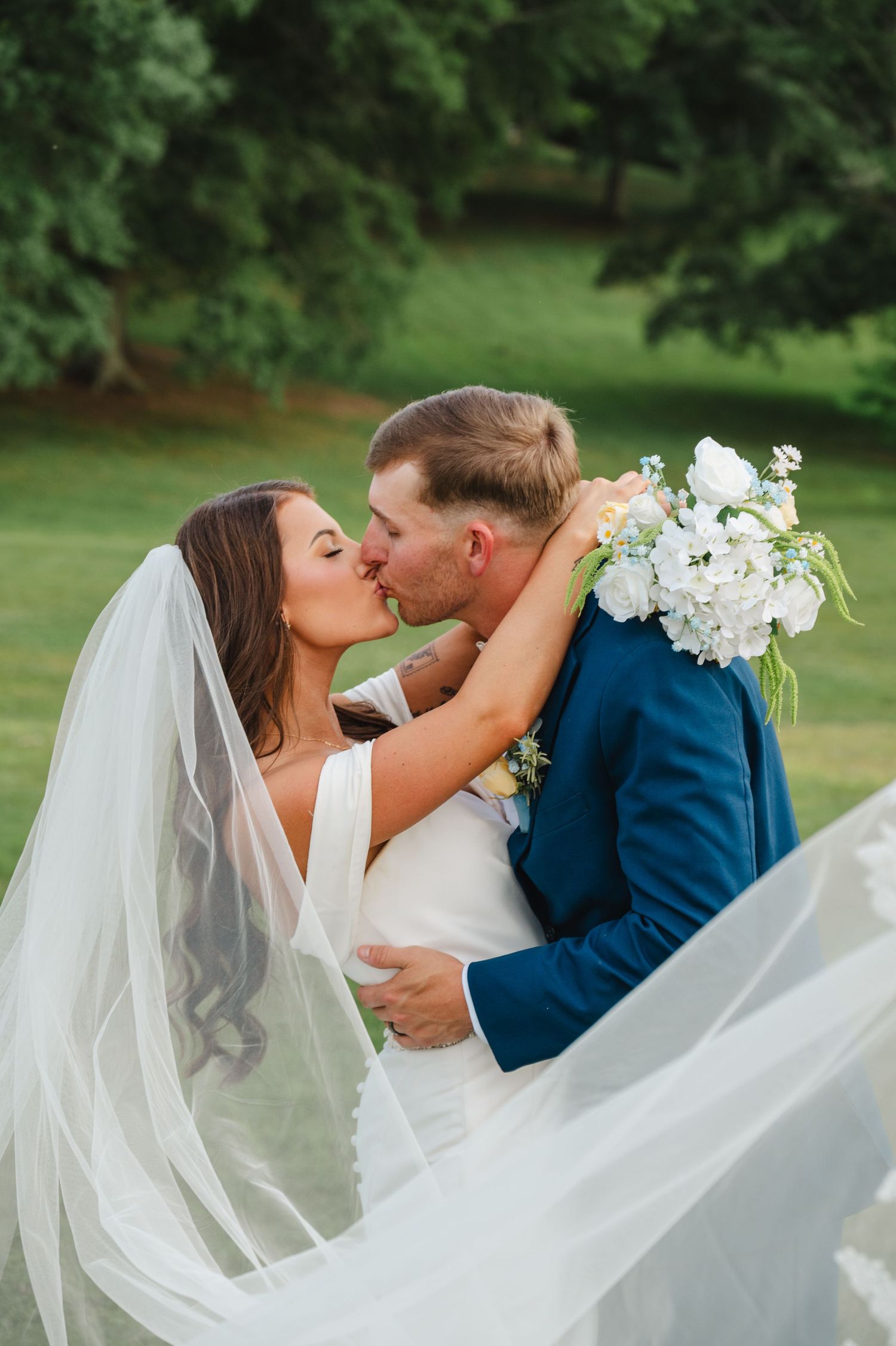 Bride and groom share a romantic kiss outdoors, with flowing white veil and bridal bouquet of white flowers.