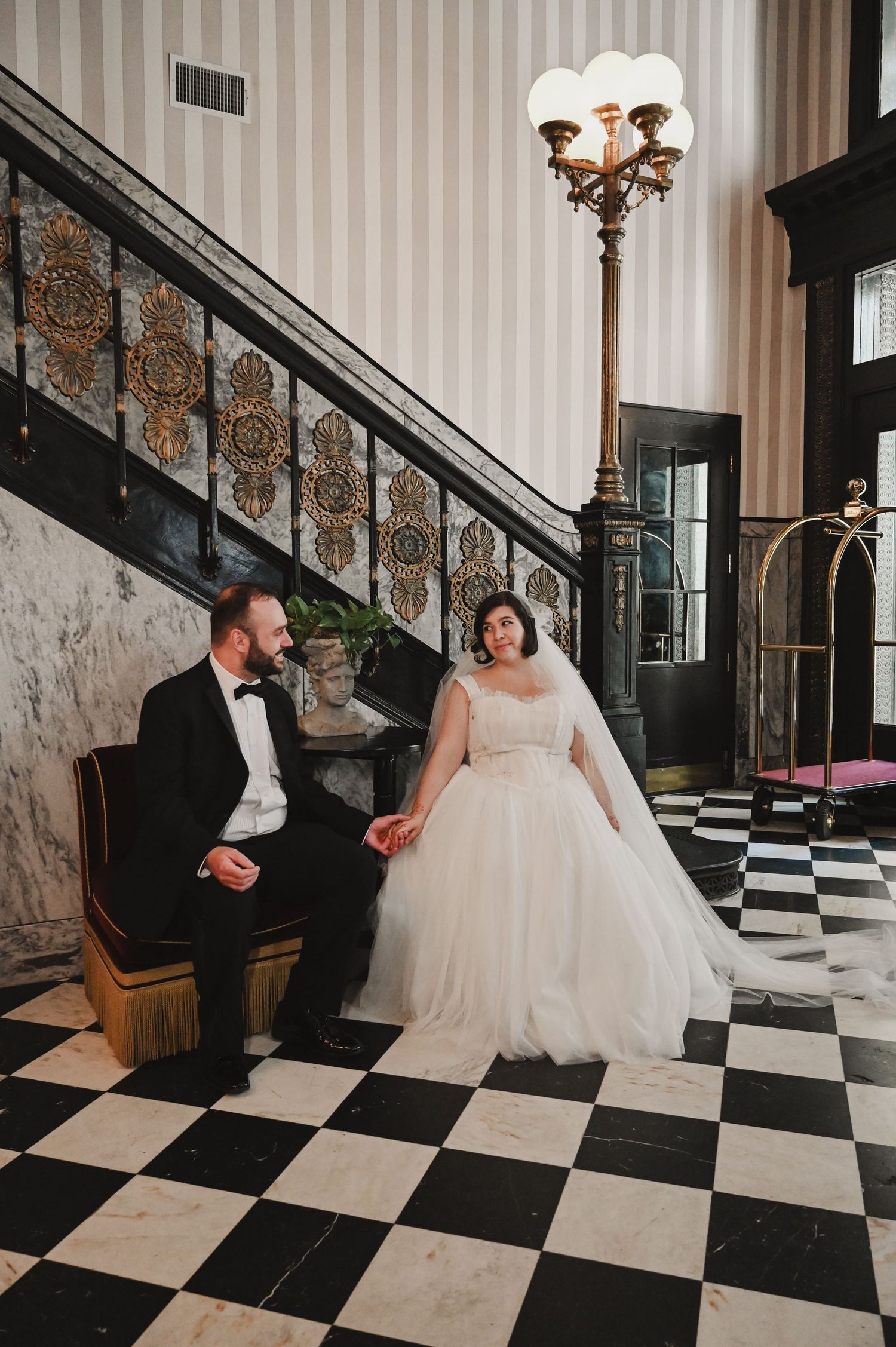 Bride and groom hold hands on black and white checkered floor by ornate staircase in elegant hotel lobby.
