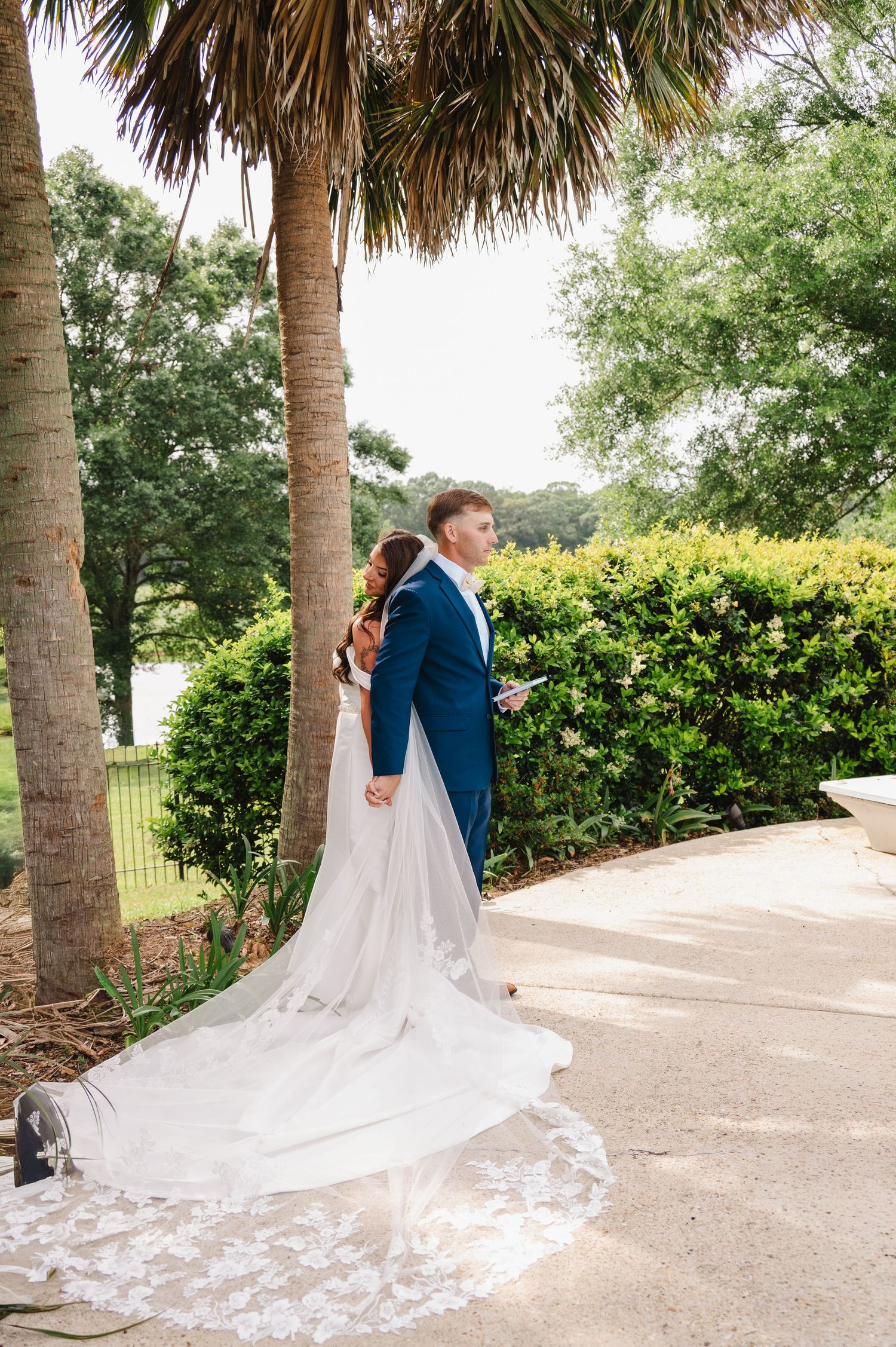 Bride in lace gown and groom in navy suit share a romantic moment among palm trees and lush greenery.