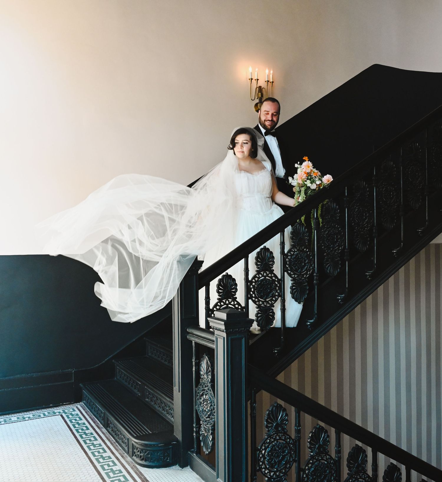Bride and groom pose elegantly on a grand staircase, her veil flowing, holding a bouquet at a classic wedding venue.