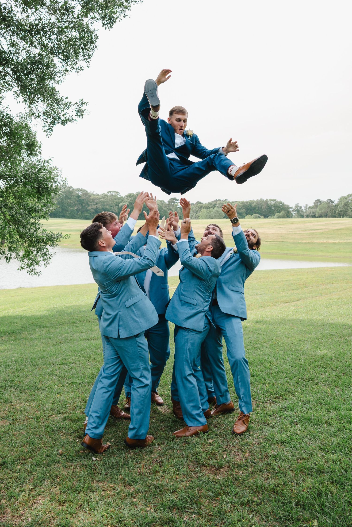 Groomsmen in light blue suits tossing the groom into the air outdoors near a lake during a wedding celebration.