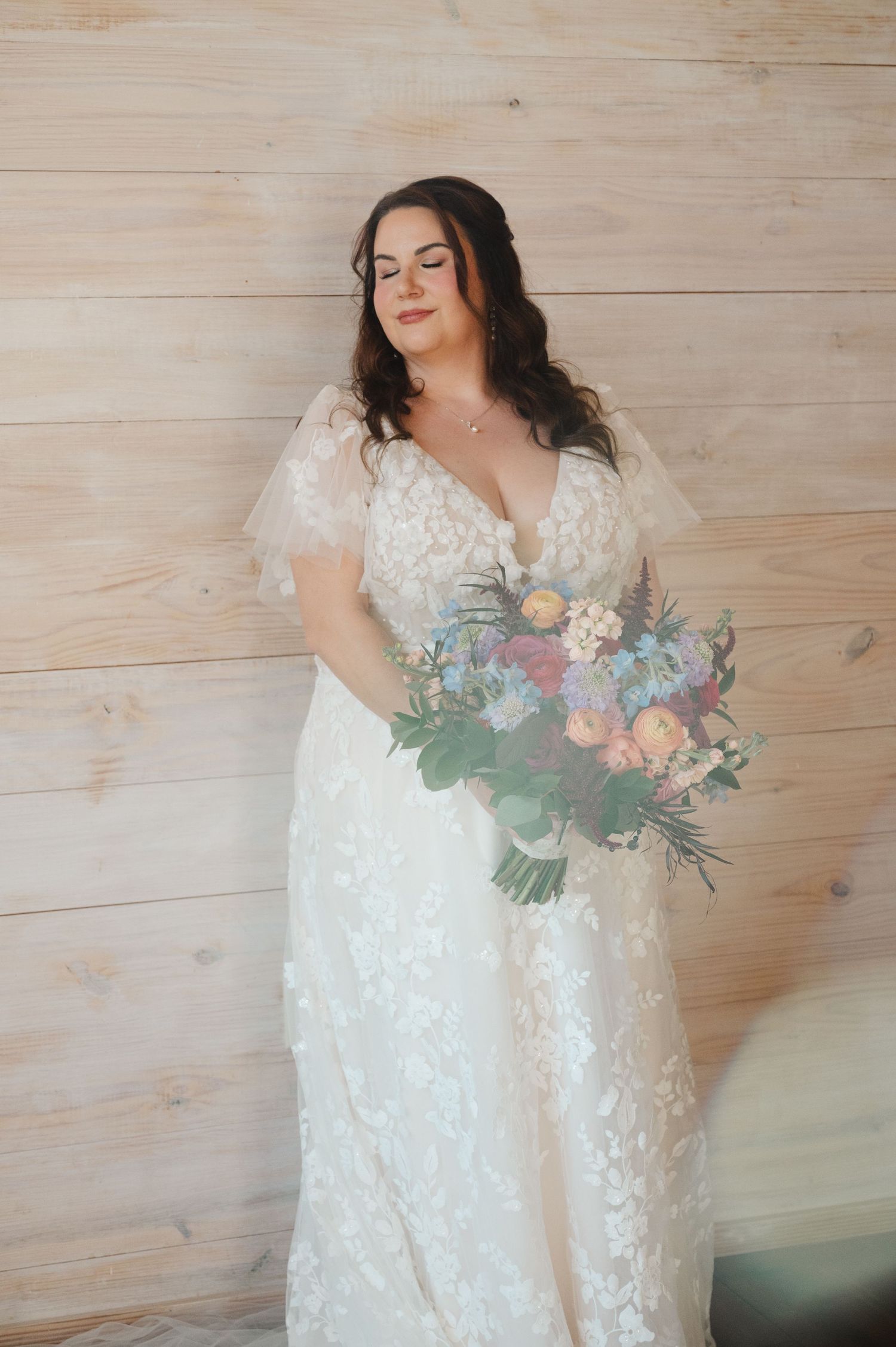 Bride in lace flutter-sleeve wedding gown holding pastel floral bouquet, posing against light wood panel wall.