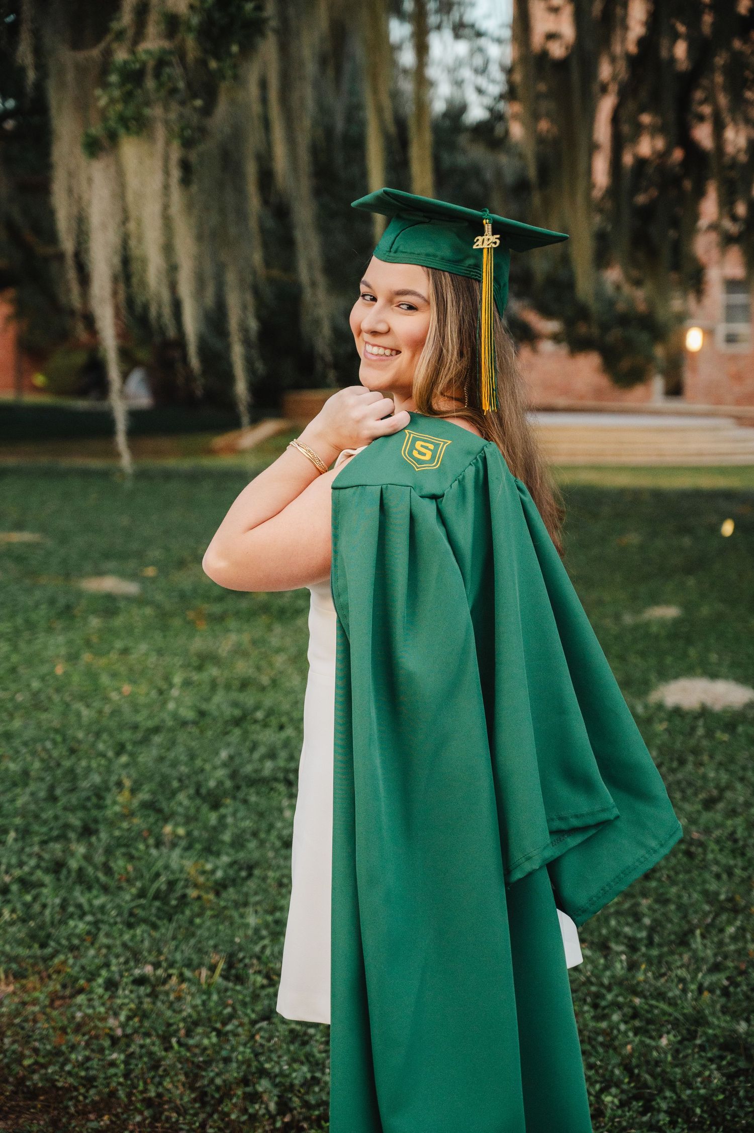 Smiling graduate in green cap and gown poses outdoors showing school emblem, with lush trees in background.