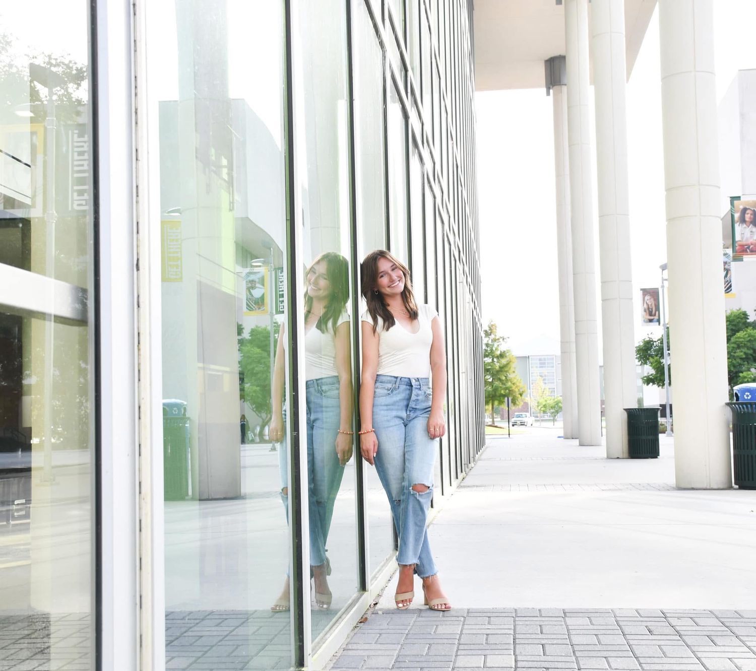 Young woman in white top and ripped jeans posing beside a reflective glass building.