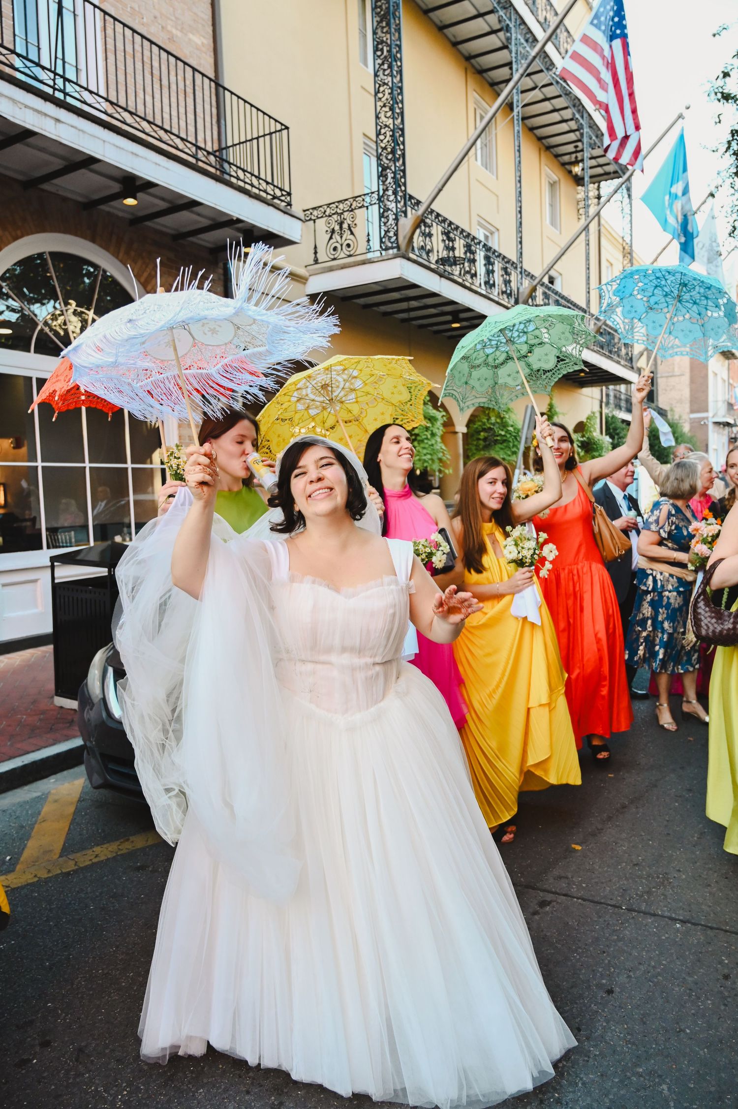 Bride in white gown leads colorful wedding parade with bridesmaids in rainbow dresses holding parasols on a sunny street.