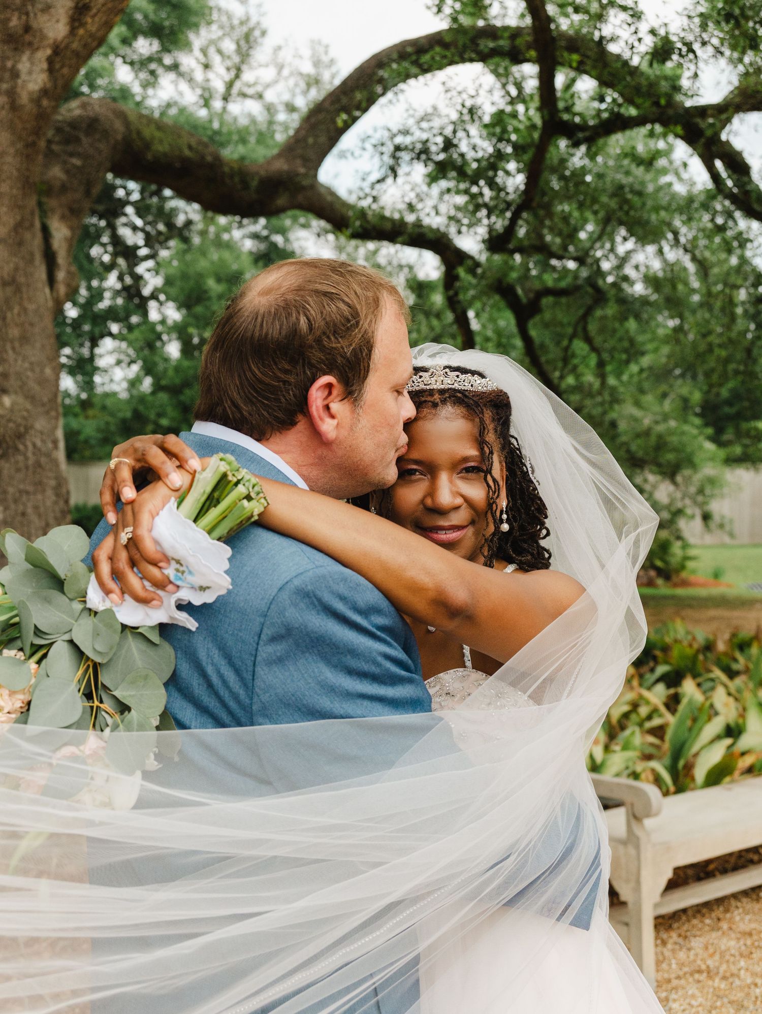 Bride and groom share a romantic embrace outdoors, with the bride's veil flowing and holding a white floral bouquet.