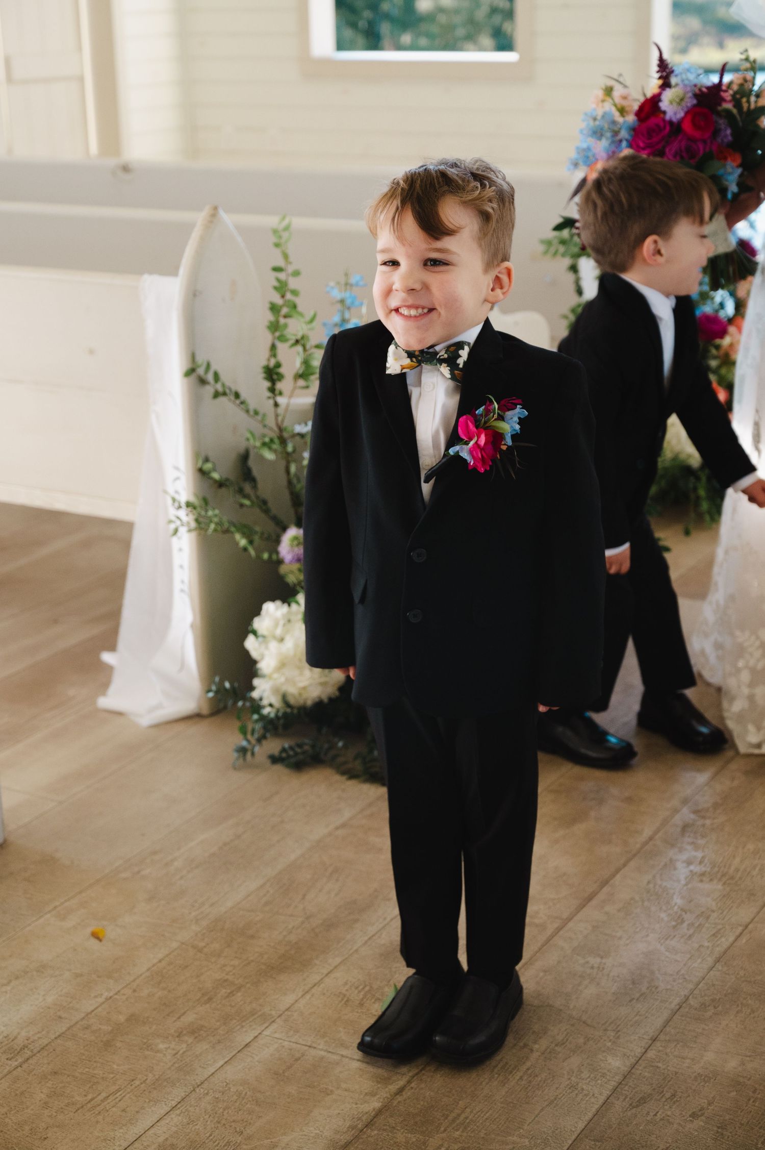 Young ring bearer in black tuxedo with floral boutonniere smiles during wedding ceremony in white chapel.