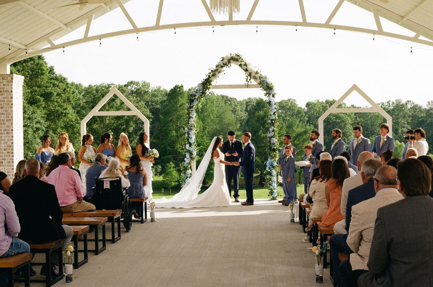 Bride and groom exchange vows under floral arch at outdoor wedding ceremony with seated guests.