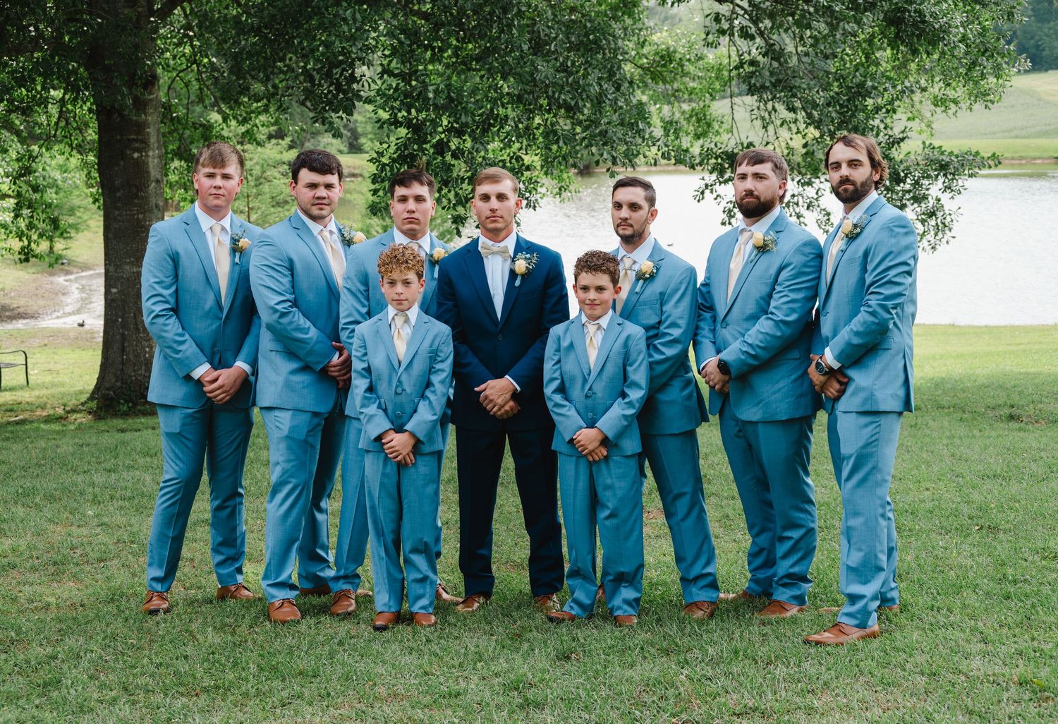 Groomsmen and groom posing in matching light blue suits with boutonnieres on green lawn near a lake.
