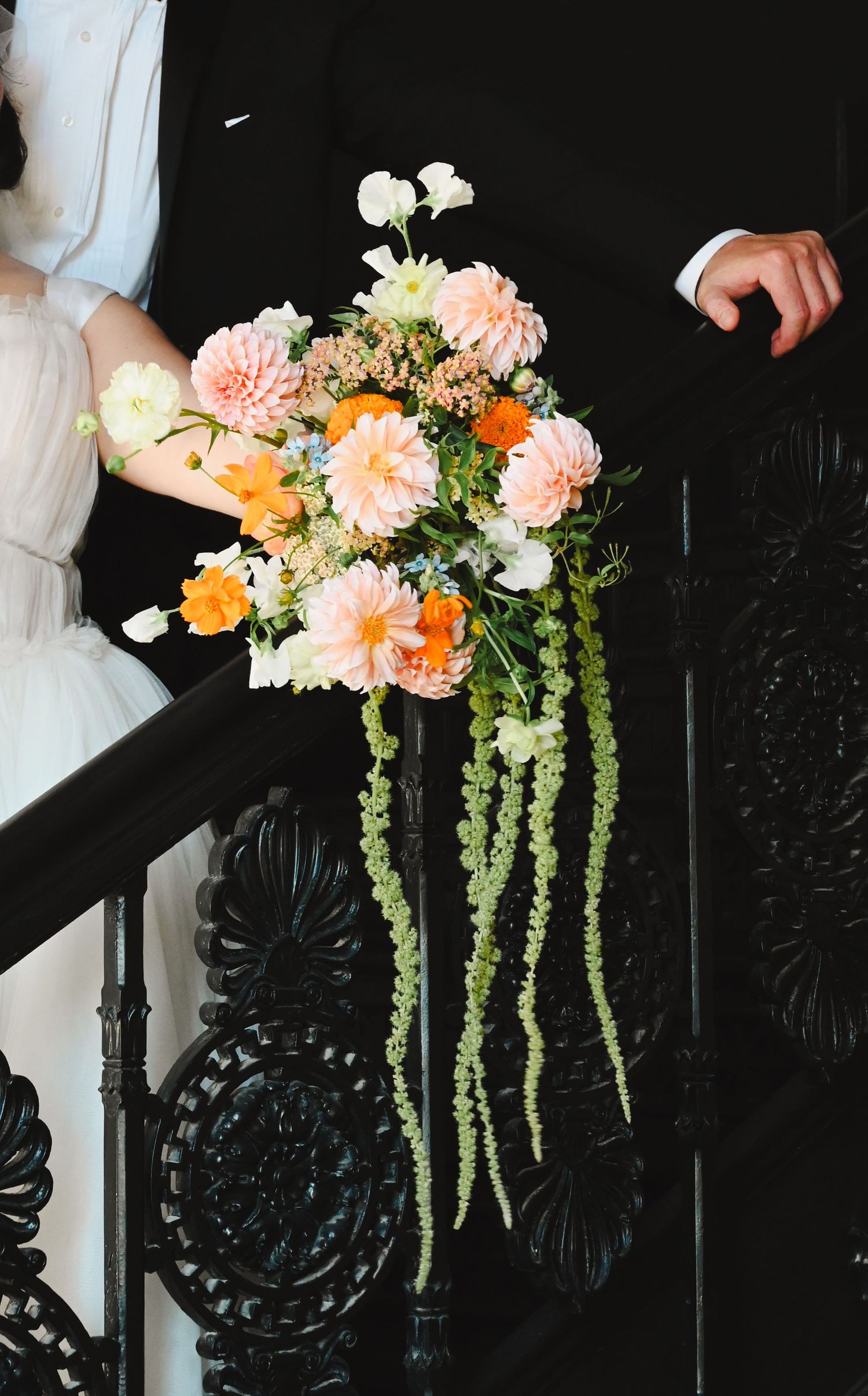 Bride holding a cascading wedding bouquet with peach dahlias, orange blooms, and trailing greenery on ornate black staircase.