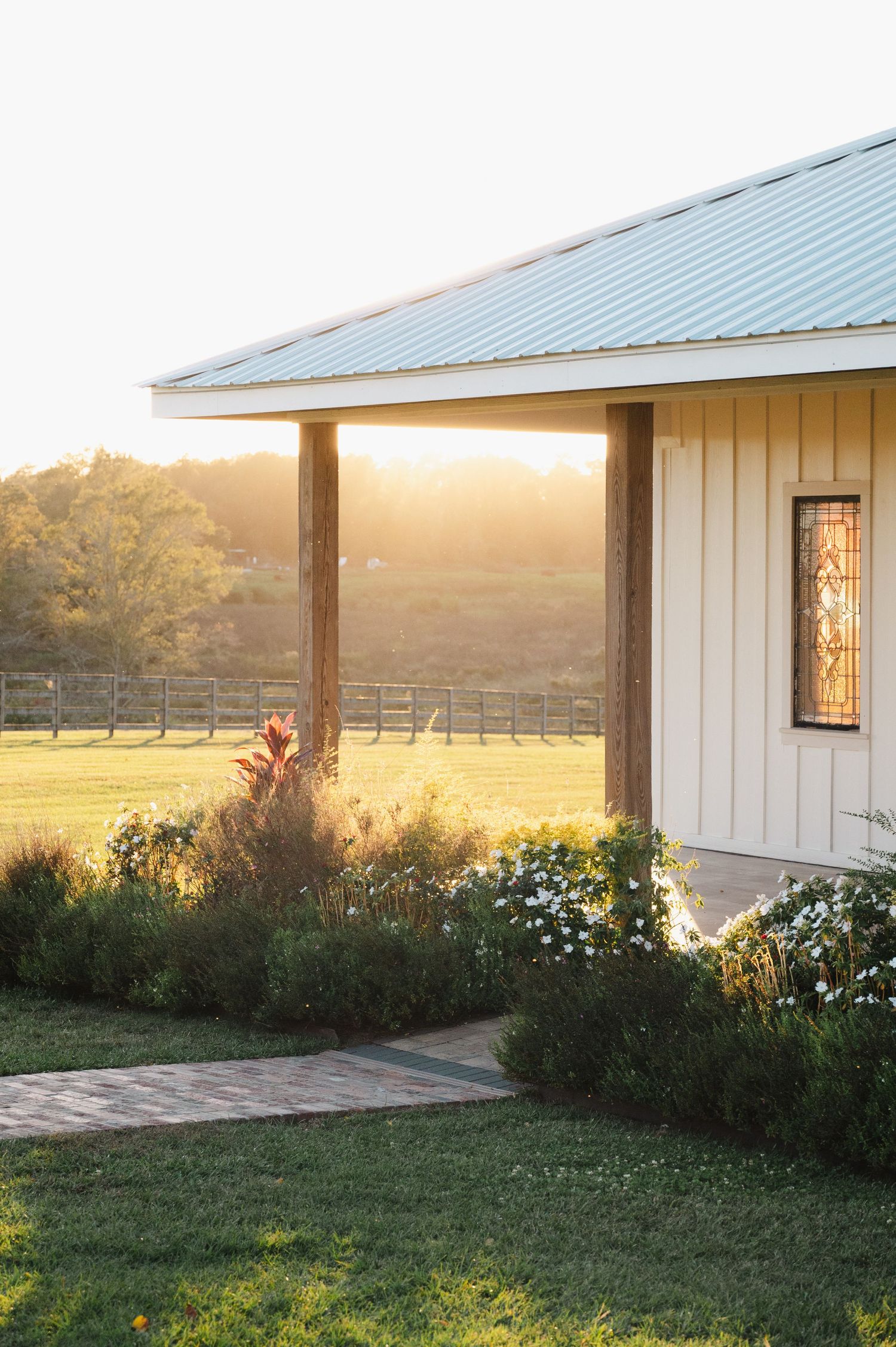 White farmhouse with metal roof at golden hour, surrounded by lush gardens and pastoral fence line at sunset.