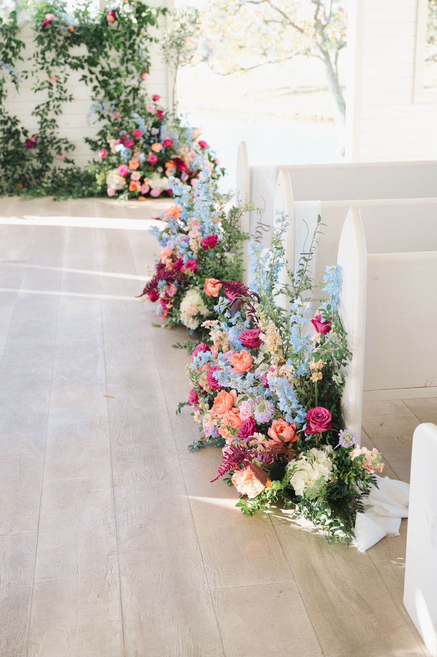Colorful wedding aisle flowers with pink, blue, and peach blooms lining white chapel pews on hardwood floors.