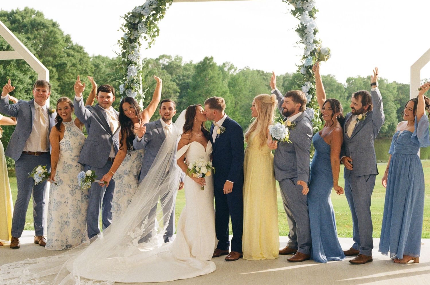Wedding party celebrates with raised arms as bride and groom kiss under a floral arch outdoors.