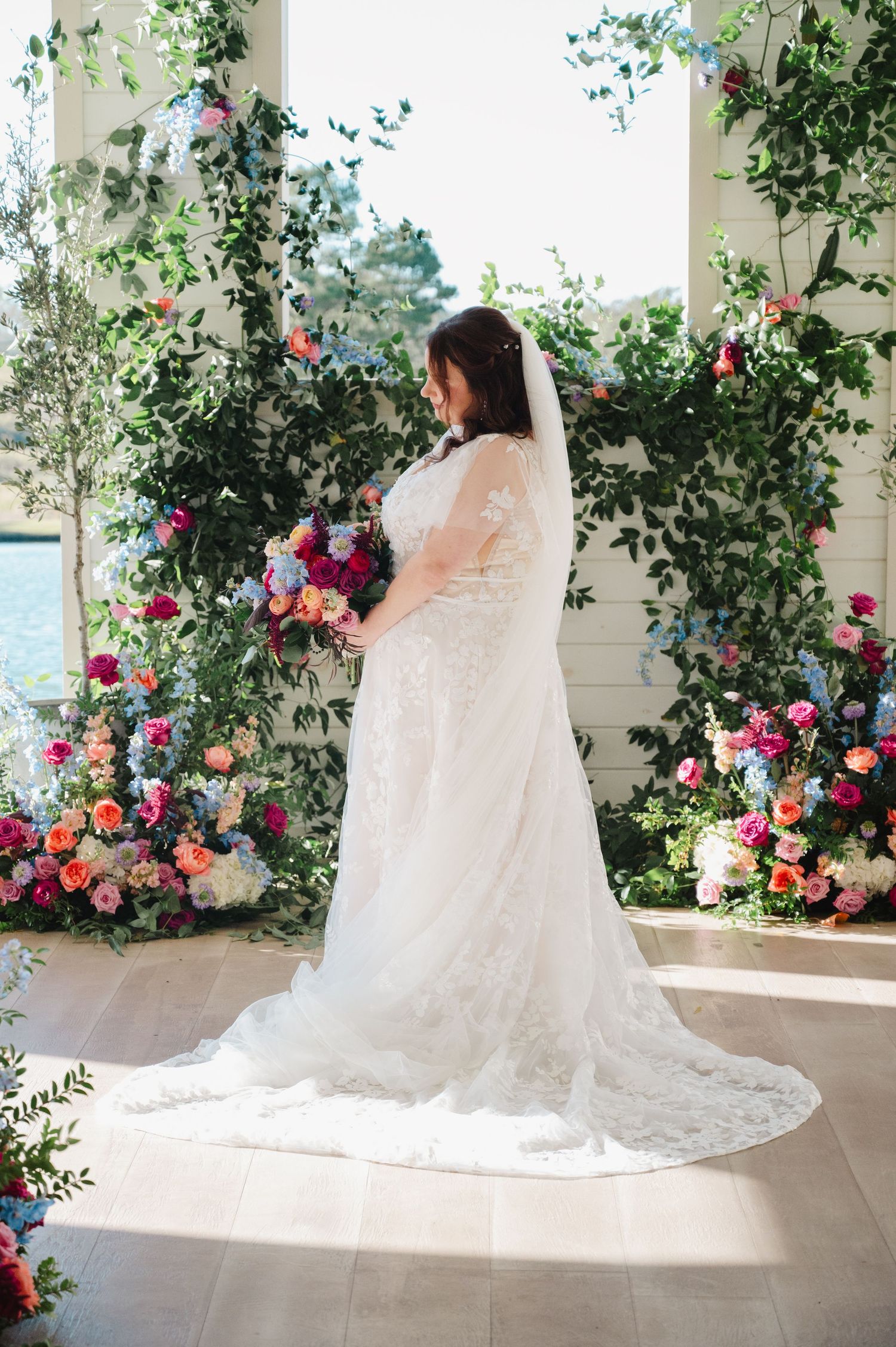 Bride in lace gown with veil holding colorful bouquet before floral arch with greenery and blooms.